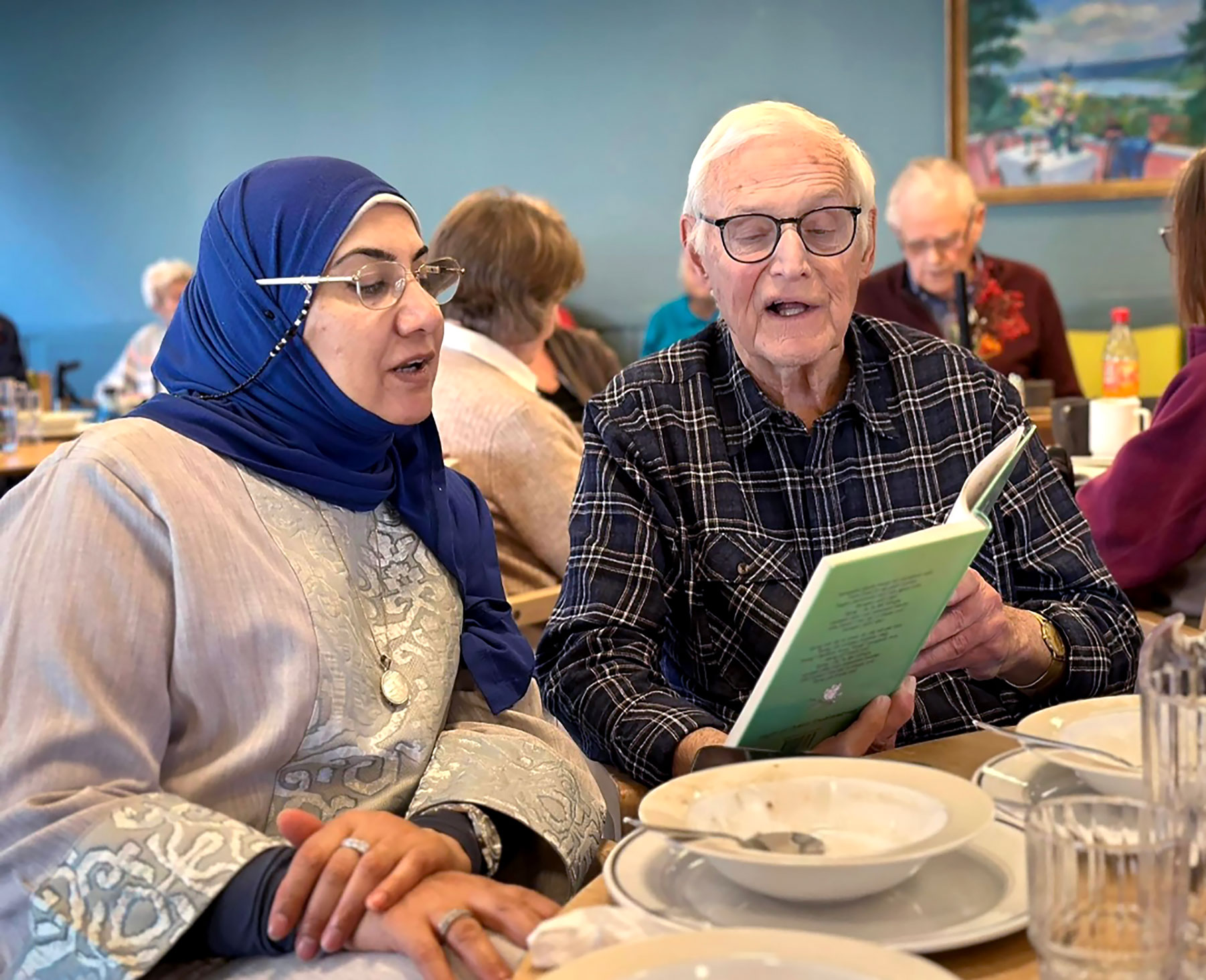 Foto: Kvinne i 40-årene med hijab og briller sitter ved siden av en mann i 80-årene og synger. Han holder opp en grønn sangbok for dem.