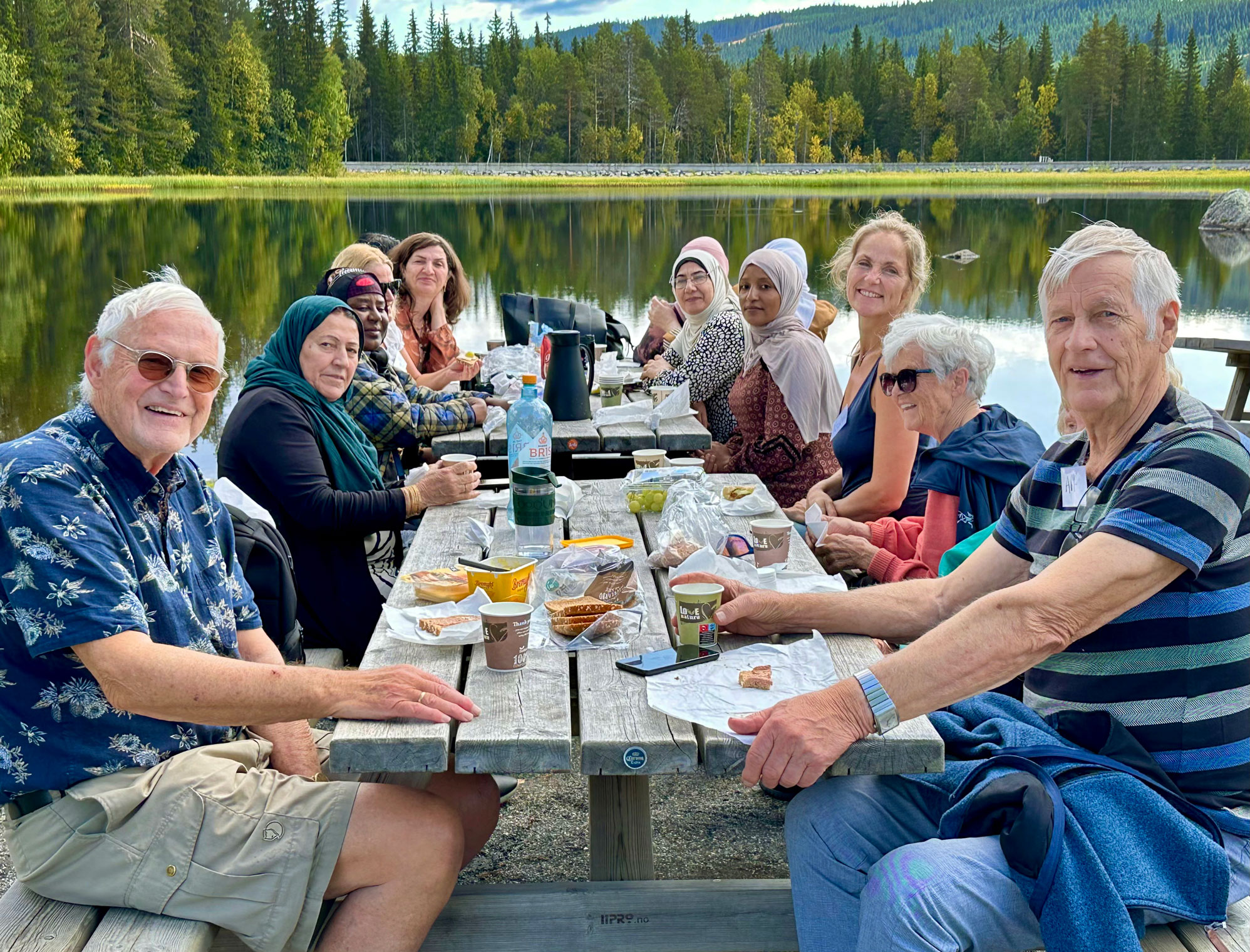 Foto: Rundt to bord og benker i tre sitter to eldre herrer og en dame sammen med flere kvinner med og uten hijab. De sitter ved et vann ute i naturen og spiser niste.