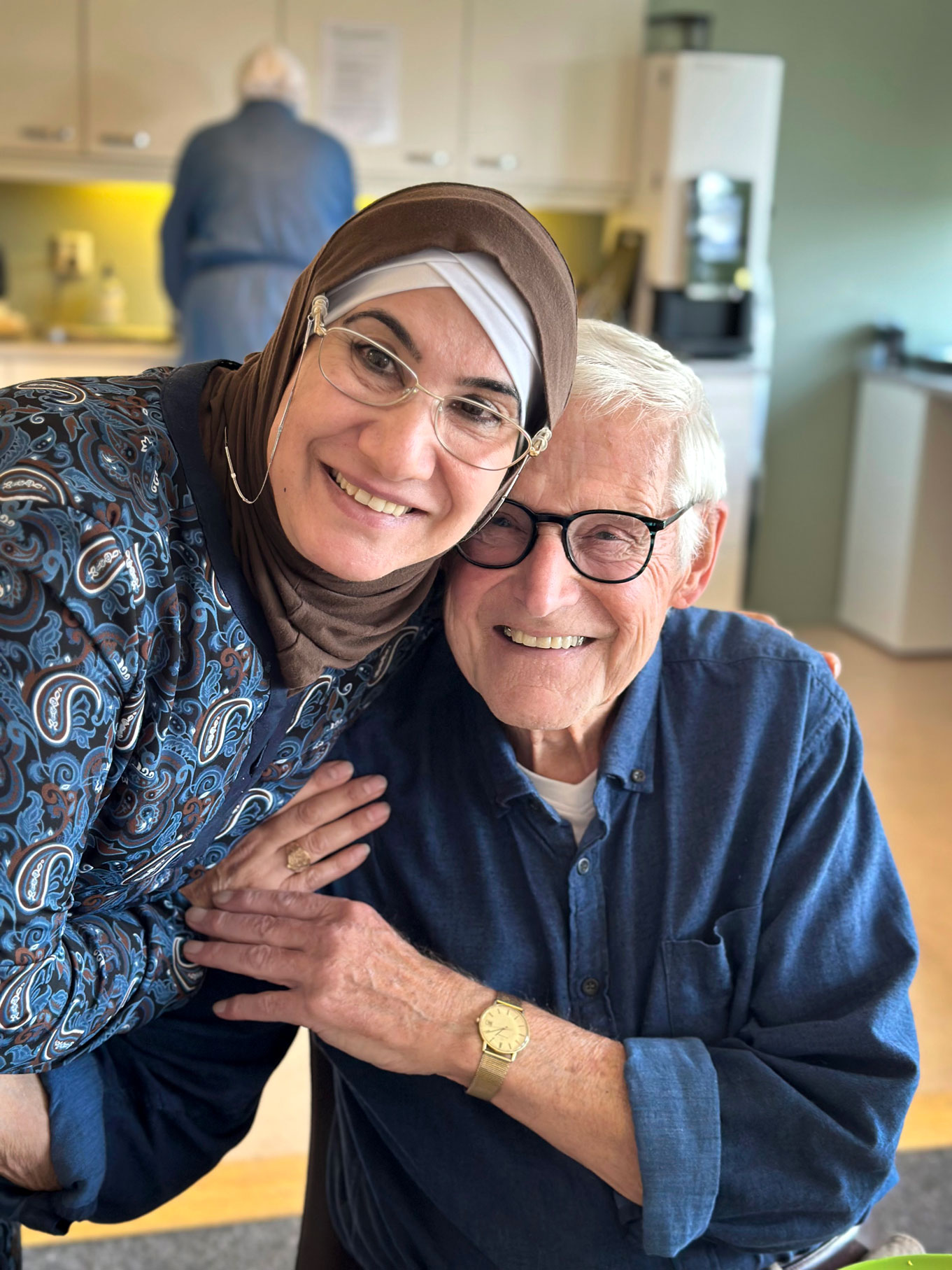Foto: Kvinne med brun hijab og briller klemmer en eldre mann med grått hår og briller i en brun skjorte.