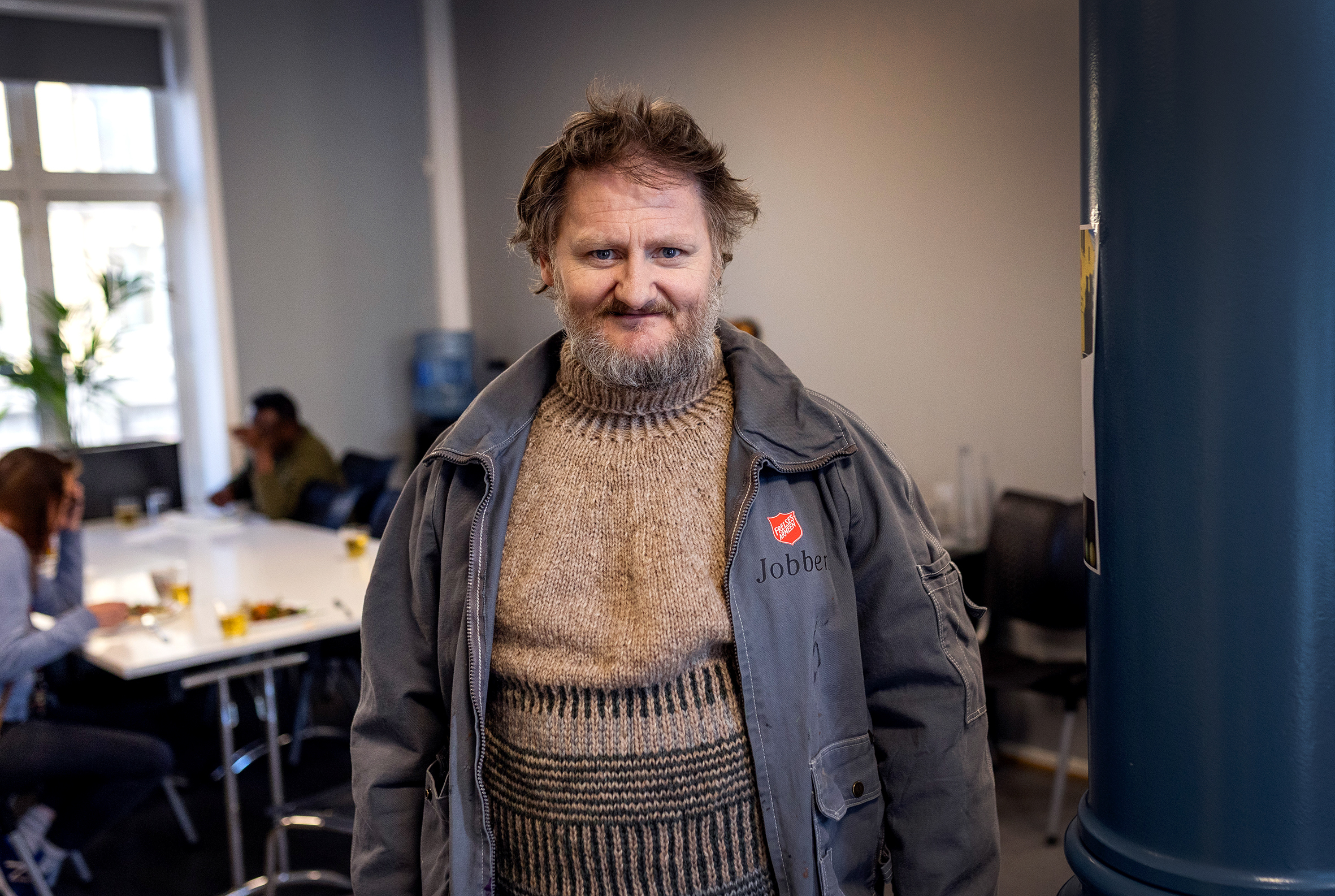 Foto: Mann med tjukk genser under en arbeidsjakke. Han har mye hår og skjegg og smiler mot kamera.