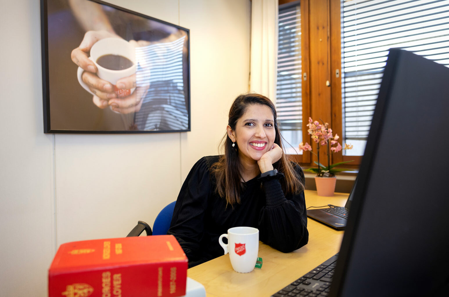 Smilende kvinne sitter foran en PC ved et skrivebord. På pulten ligger en tjukk, rød bok med Norges lover og det står en kopp med logo fra Frelsesarmeen på.