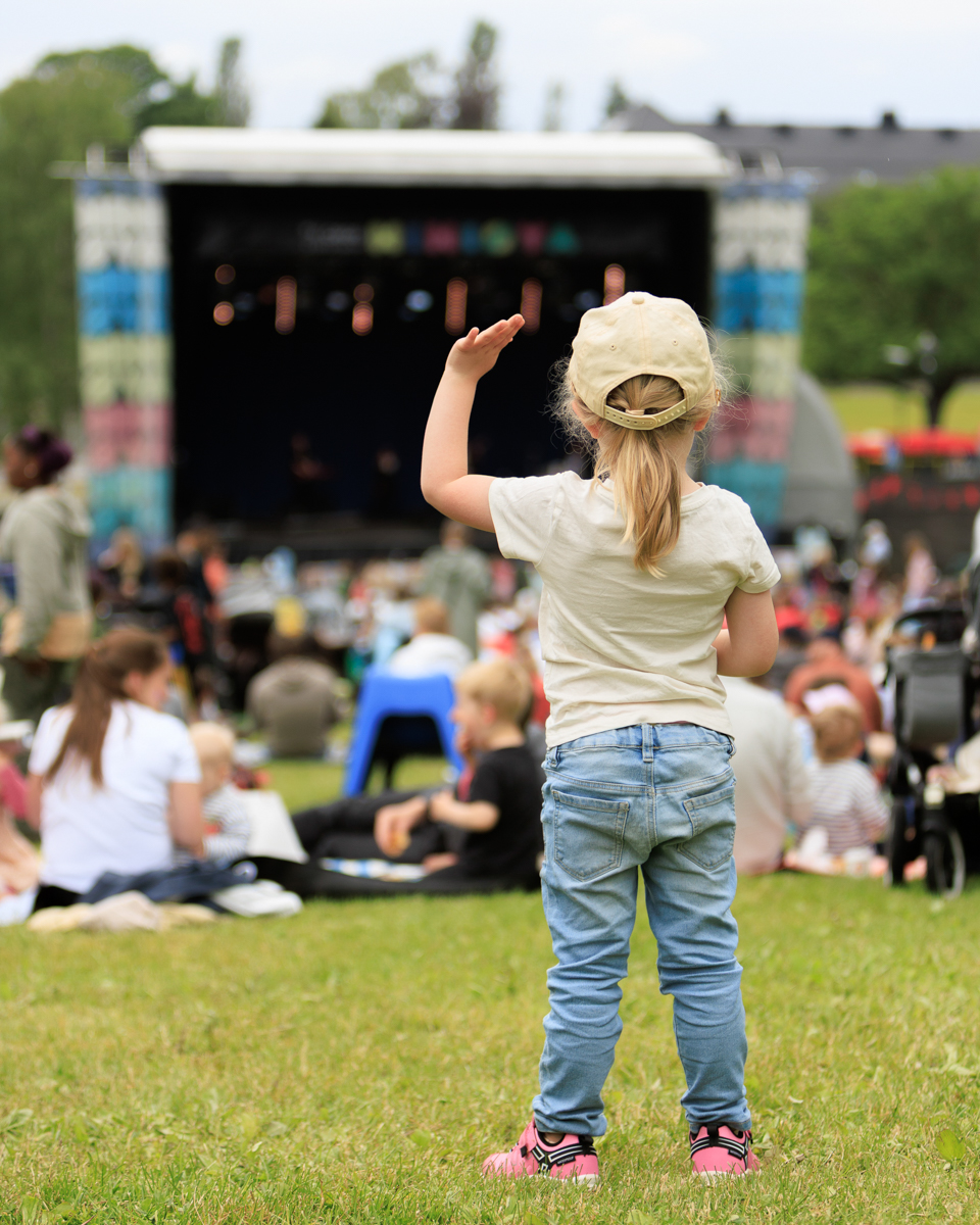 Foto: Ei lita jente i olabukser, hvit t-skjorte og caps står med ryggen til, med en hånd i været. Foran henne sees en scene og mange mennesker som sitter på gresset, de er uskarpe i forgrunnen.
