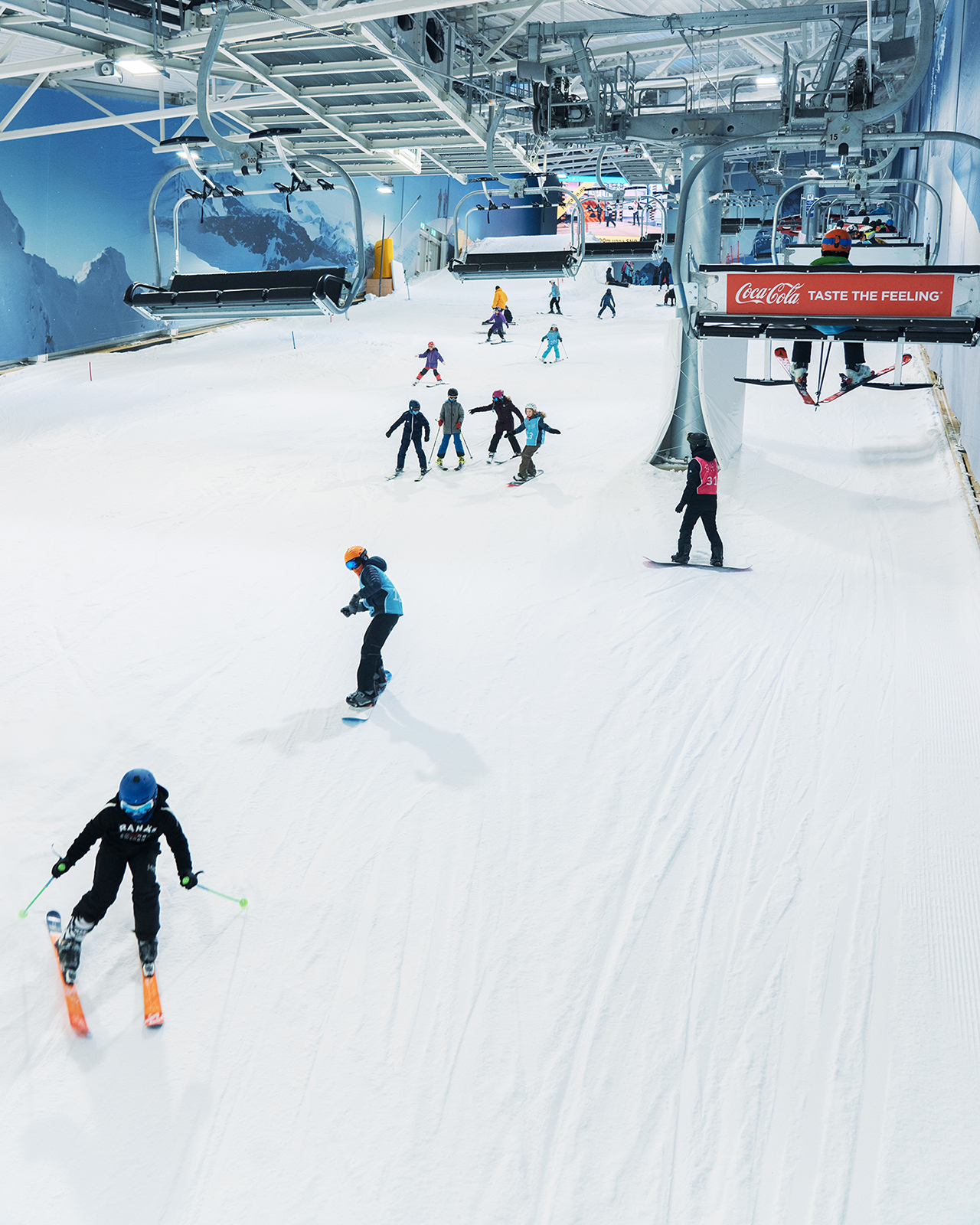 Foto: En innendørs slalombakke, mange personer på ski og snowboard er på vei ned bakken, de sees på avstand. På venstre side er det en skiheis.