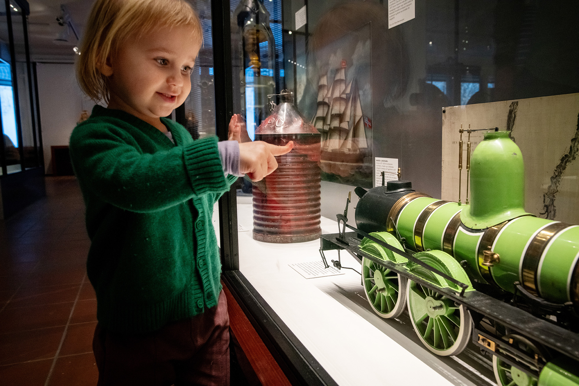 Foto: Ei lita jente med halvlangt lyst hår og grønn strikket genser står og smiler og ser inn i en monter med en modell av et tog. Hun trykker fingeren mot glasset i monteren.