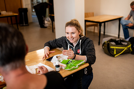 Foto: Ung kvinne sitter ned på huk ved et bord i et stort rom. Smiler og snakker til en mann. Det er flere rundt om i rommet, men en ser ingen andre ansikter.