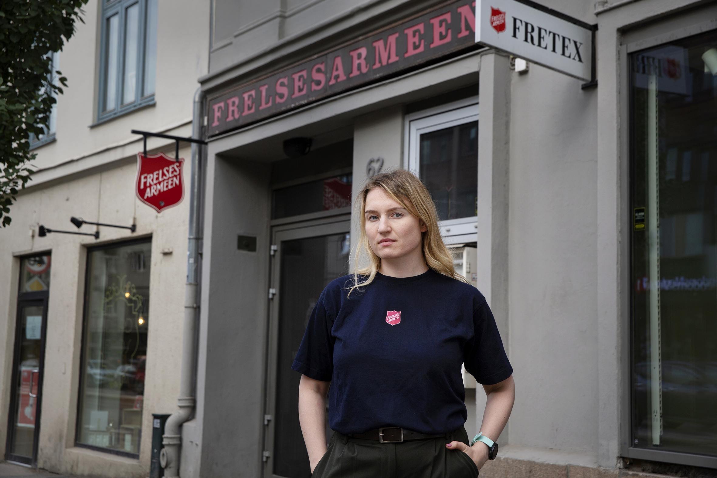 Foto: Kvinne i blå T-skjorte med Frelsesarmeens logo på brystet. Hun står utenfor en gammel bygård i betong med skilt både til Fretex og Frelsesarmeen.
