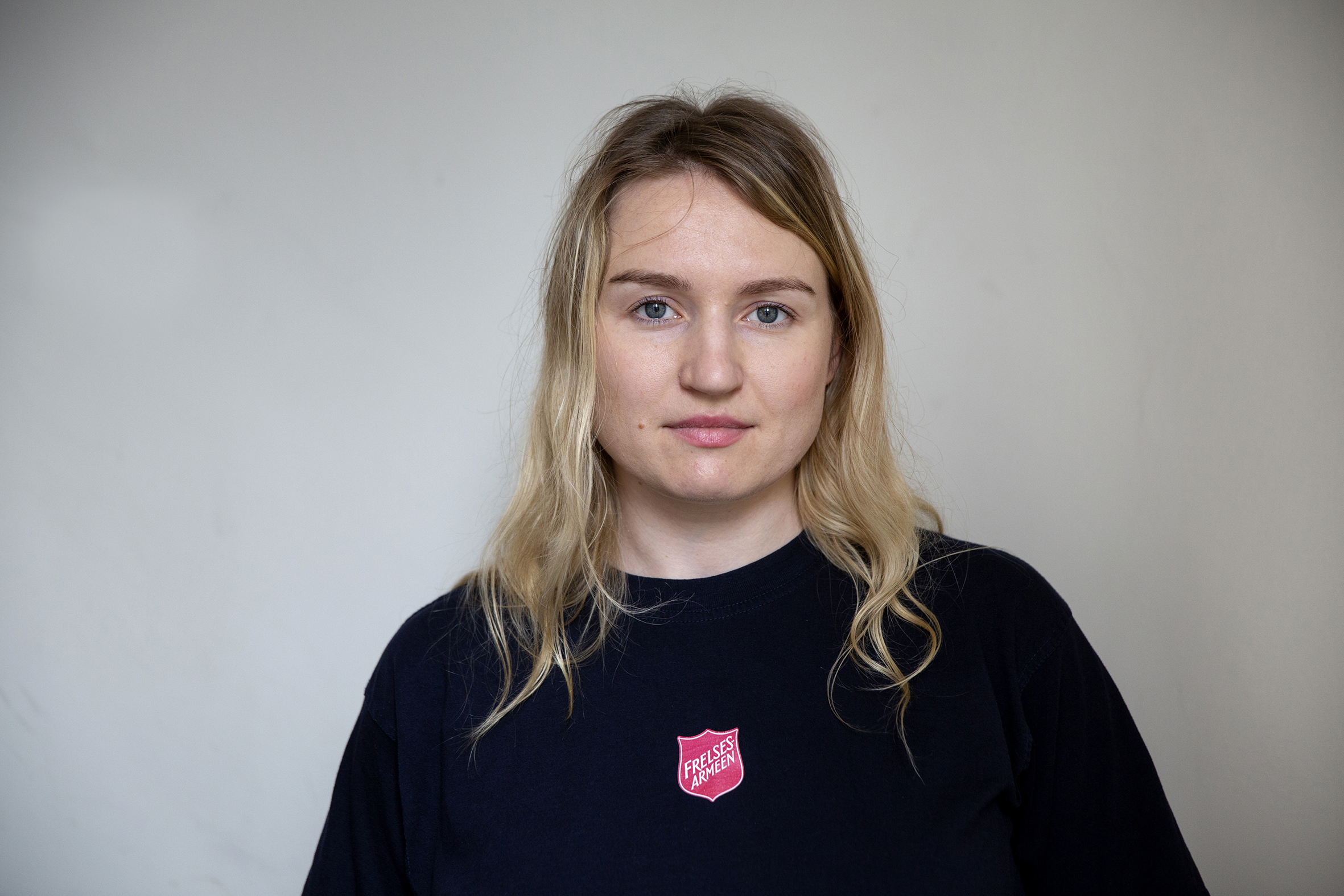 Foto: Portrett av en kvinne i 30-årene med langt, blondt hår. Hun står inn mot en hvit vegg og har på seg en blå T-skjorte med røde logo fra Frelsesarmeen.