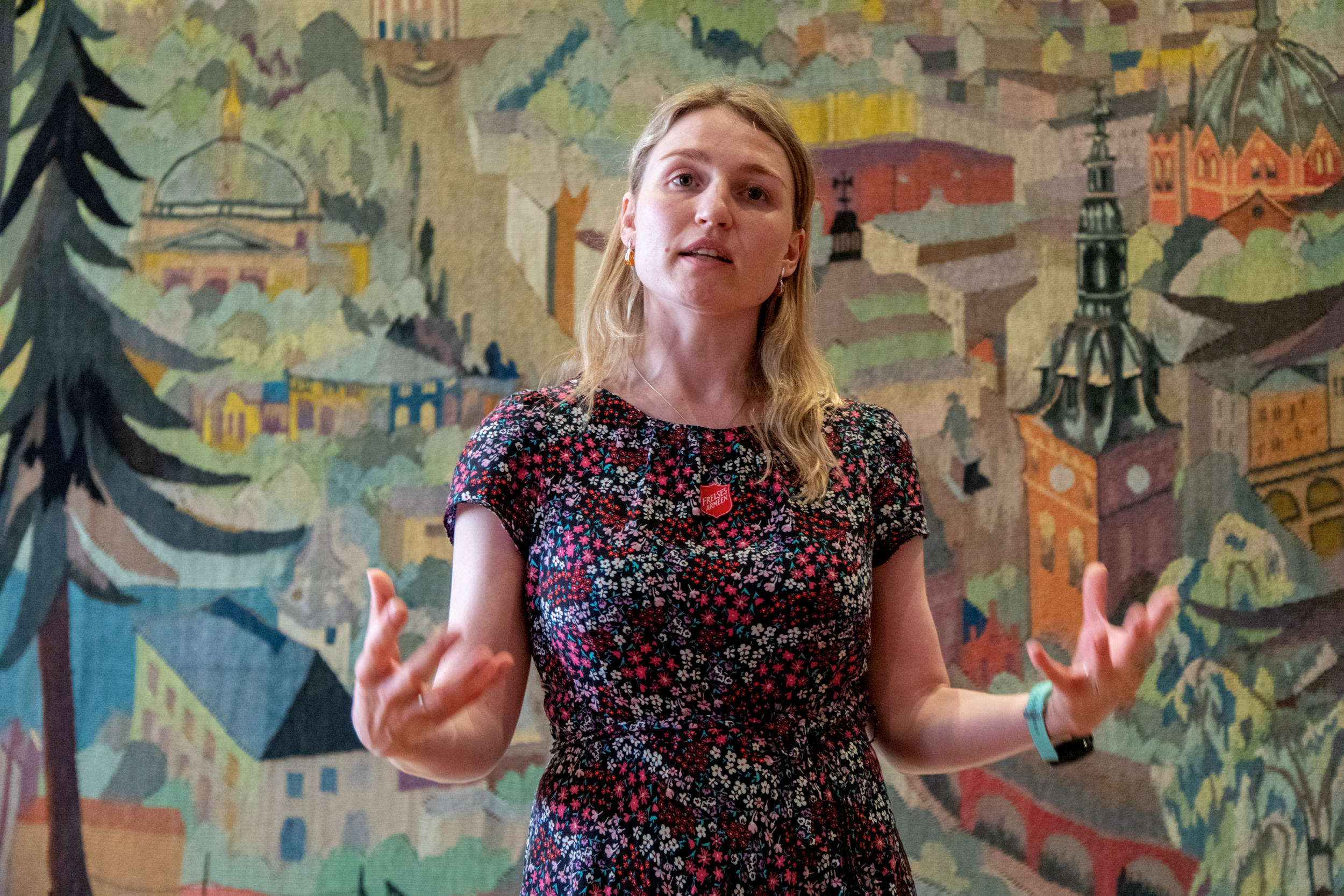 Foto: Kvinne i 30-årene i blomstrente kjole snakker. Gestikulerer med hendene. I bakgrunnen er det et fargerikt, vevd teppe med motiv fra kvartalene rundt Oslo domkirke.