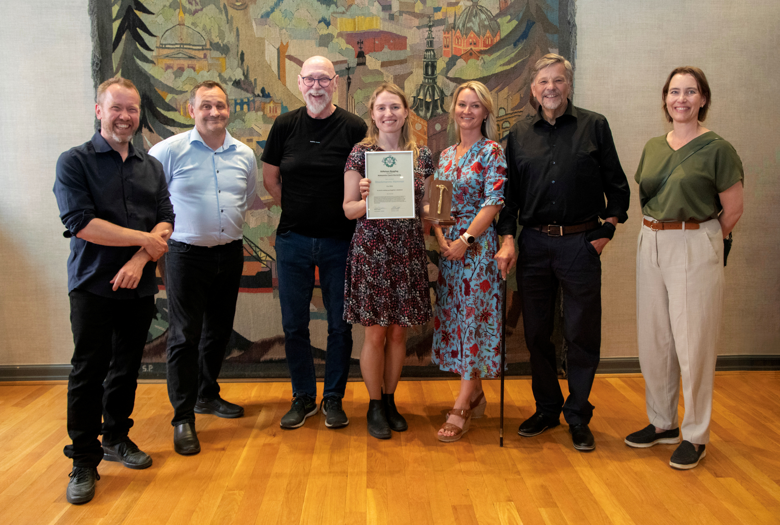 Foto: "Lagbilde" av sju mennesker oppstilt på en rekke. Fire menn og tre kvinner. Hun i midten holder opp diplomet.