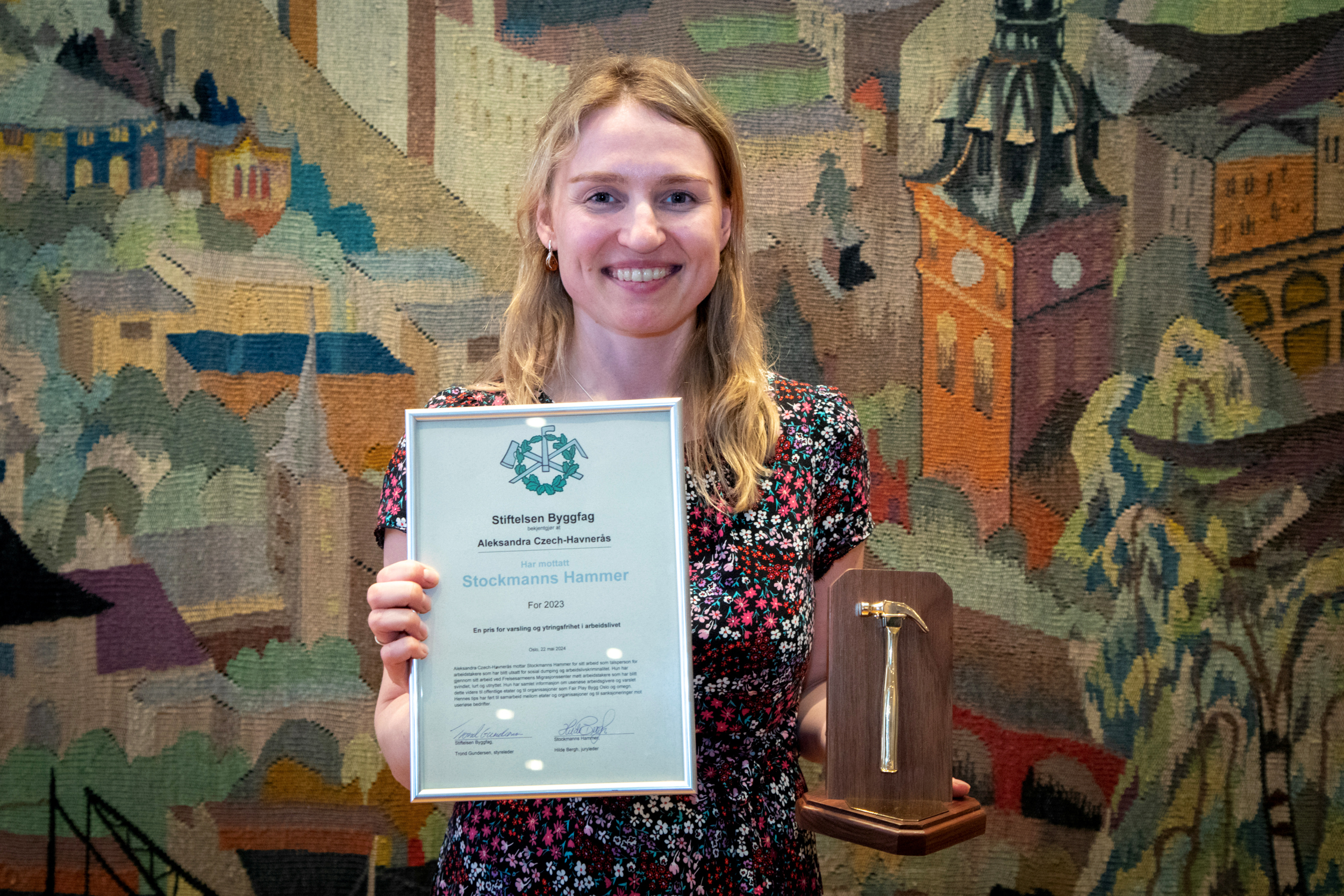 Foto: Smilende, blond kvinne i 30-årene holder opp et diplom i ramme, der det står Stockmanns hammer og hennes navn. I den andre hånden holder hun en liten gullhammer som står på et stativ i tre.