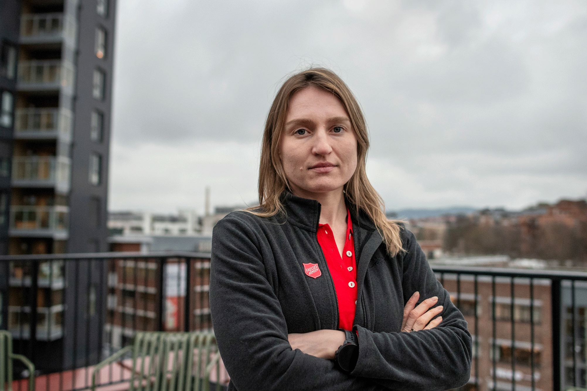 Foto: Mørkeblond kvinne i 30-årene står med armene i kors på en terrasse med rekkverk. I bakgrunnen er byen og hun har på seg et rødt skjold fra Frelsesarmeen.