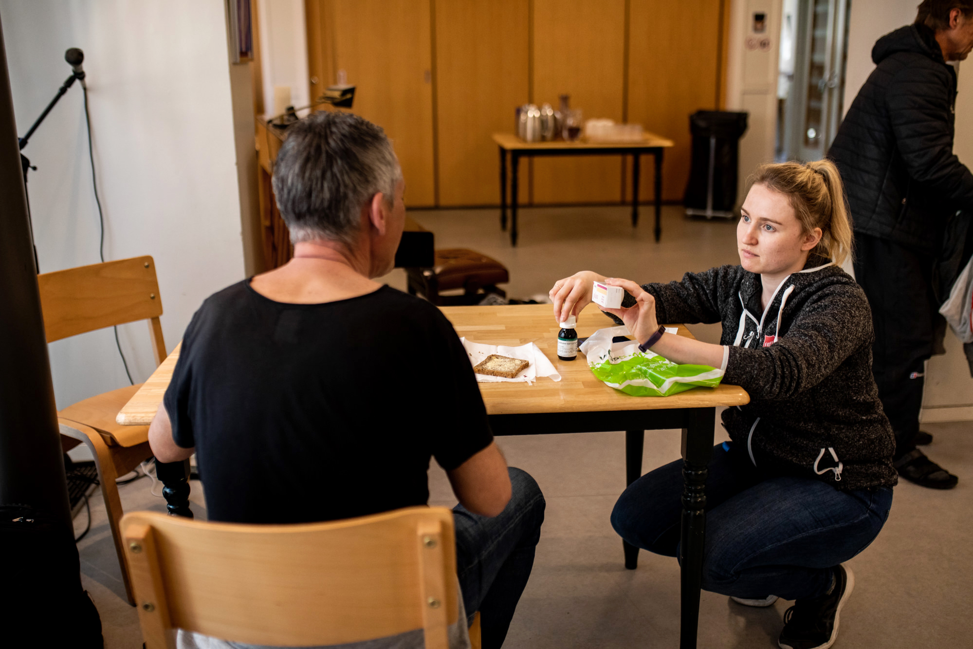 Foto: Kvinne i 30-årene med blond hestehale sitter på huk ved et bord og viser fram en flaske med medisin til en mann. han sitter på en stol med ryggen til kamera.