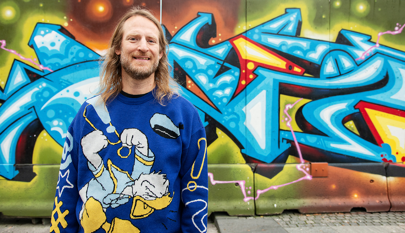 Foto: Blond mann i slutten av 40-årene står i en blå og gul genser med en and på. Han har halvlangt hår og står foran en vegg med fargerik graffiti.