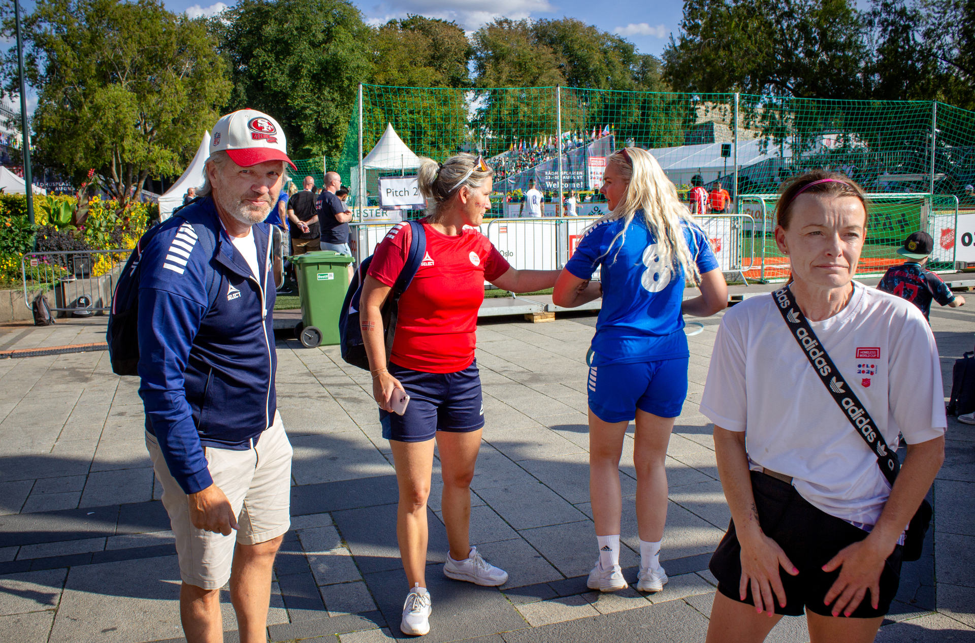 Foto: Fire personer i treningstøy, en av dem er mann. De står ute på en plass der det er fotballbinger rundt og grønne trær.