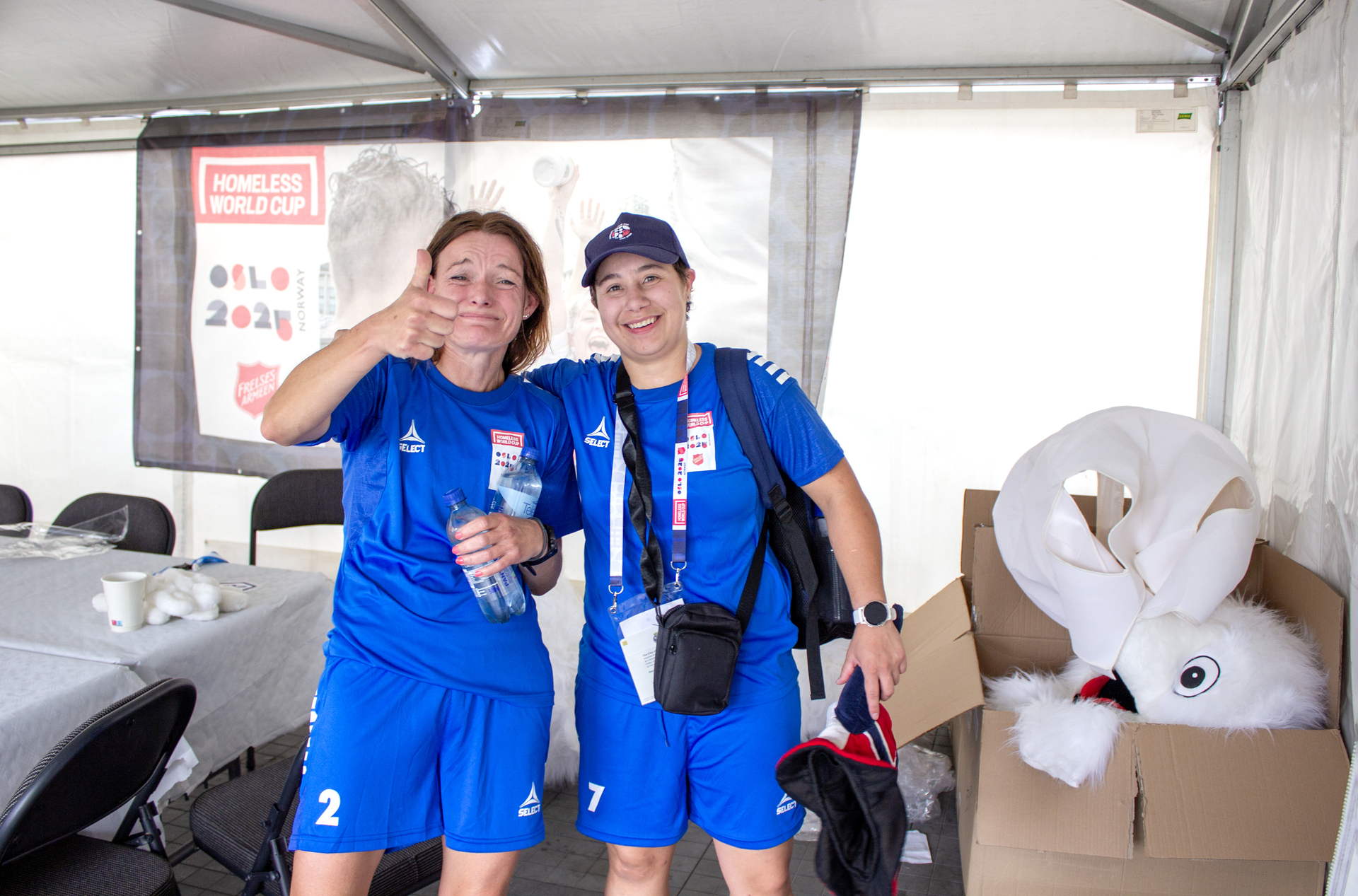 Foto: To kvinner i blå fotballdrakter står inni et telt. Den ene er ekstra svett og holder en tommel opp. I hjørnet står en eske med kostymene til to hvite isbjørn-maskoter.