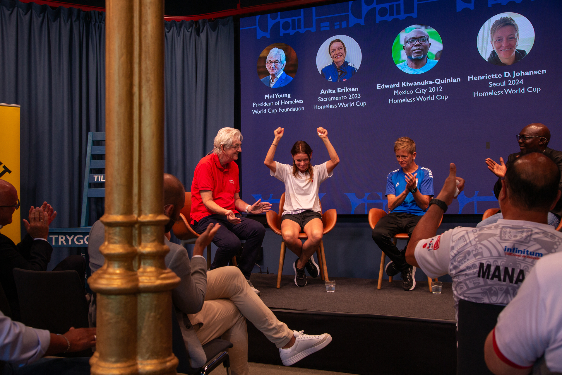 Foto: På en scene sitter fire personer på rekke, først en mann med grått hår i en rød piquet-skjorte. Deretter sitter en sped dame med mørkt hår som holder hendene opp i været. Ved siden av der igjen sitter en blond kvinne med kort hår i blå fotballtrøye og en svart mann.