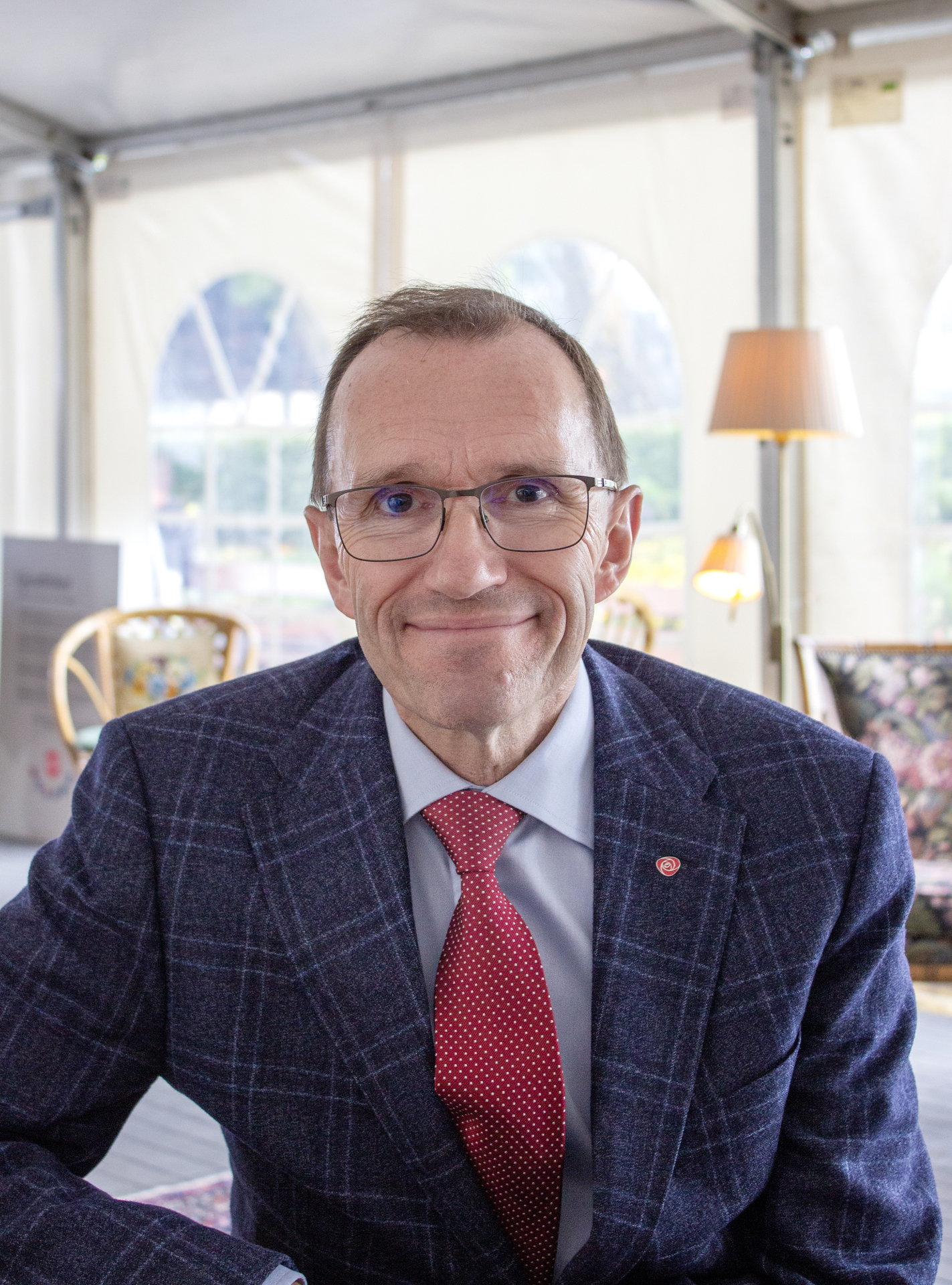 Foto: Mann i mørk dress med rødt slips og briller ser i kamera. Han sitter inne i et telt møblert med gjenbrukte, gamle møbler.