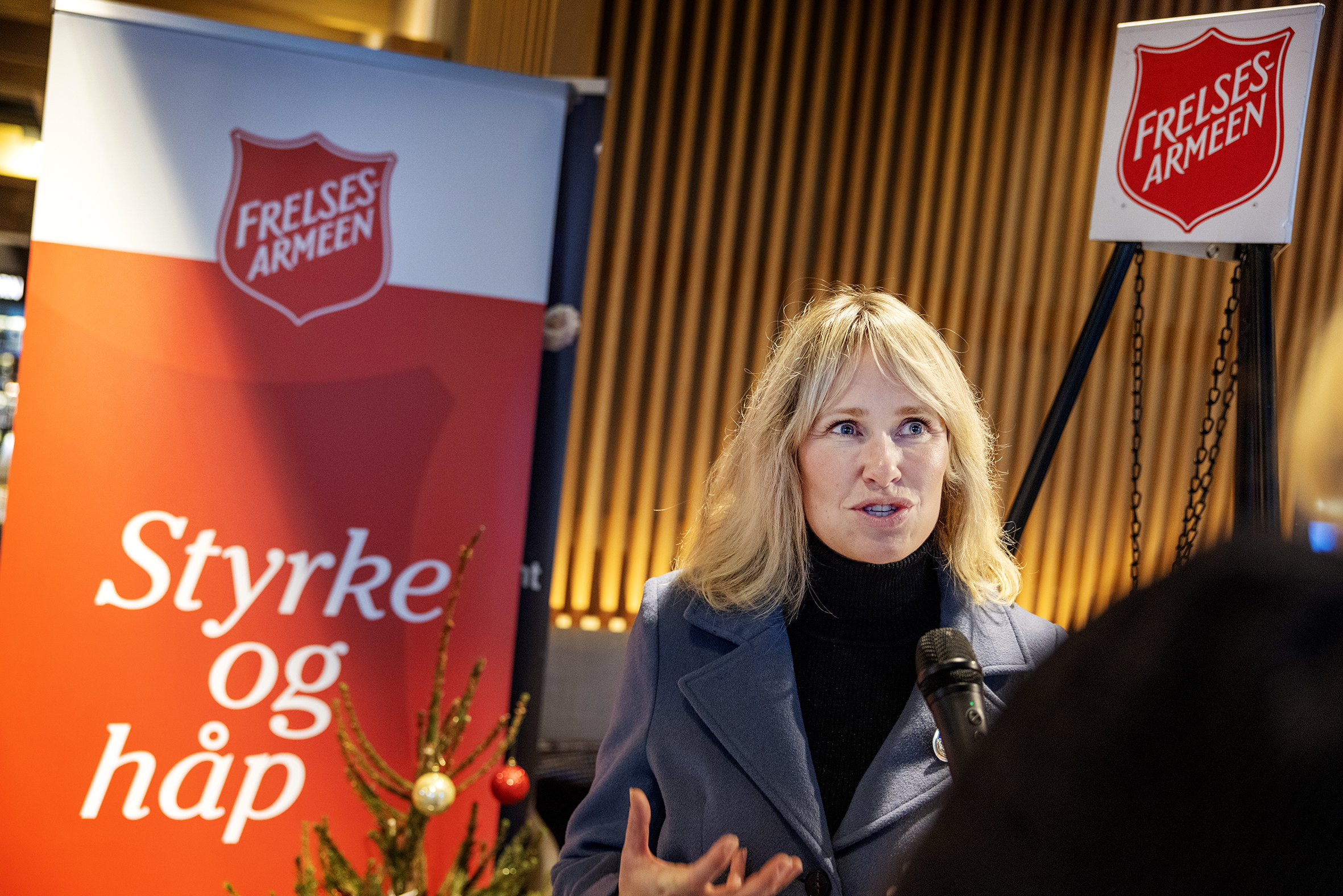 Engasjert kvinne snakker. Står foran en Frelsesarmee-plakat der det står Styrke og håp. I hjørnet står Frelsesarmeens julegryte, toppen av stativet den henger i.