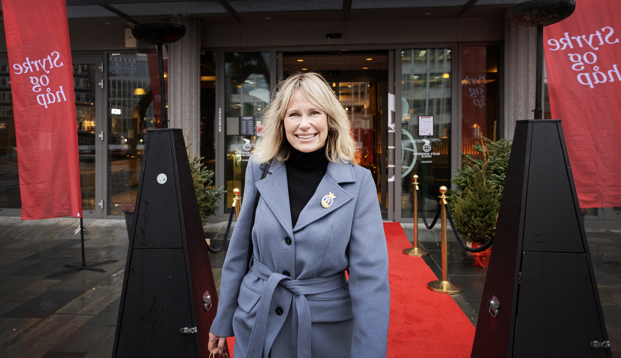 Voksen, blond kvinne står i ullkåpe ute foran en rød løper. I bakgrunnen et par røde flagg der det står Styrke for håp.