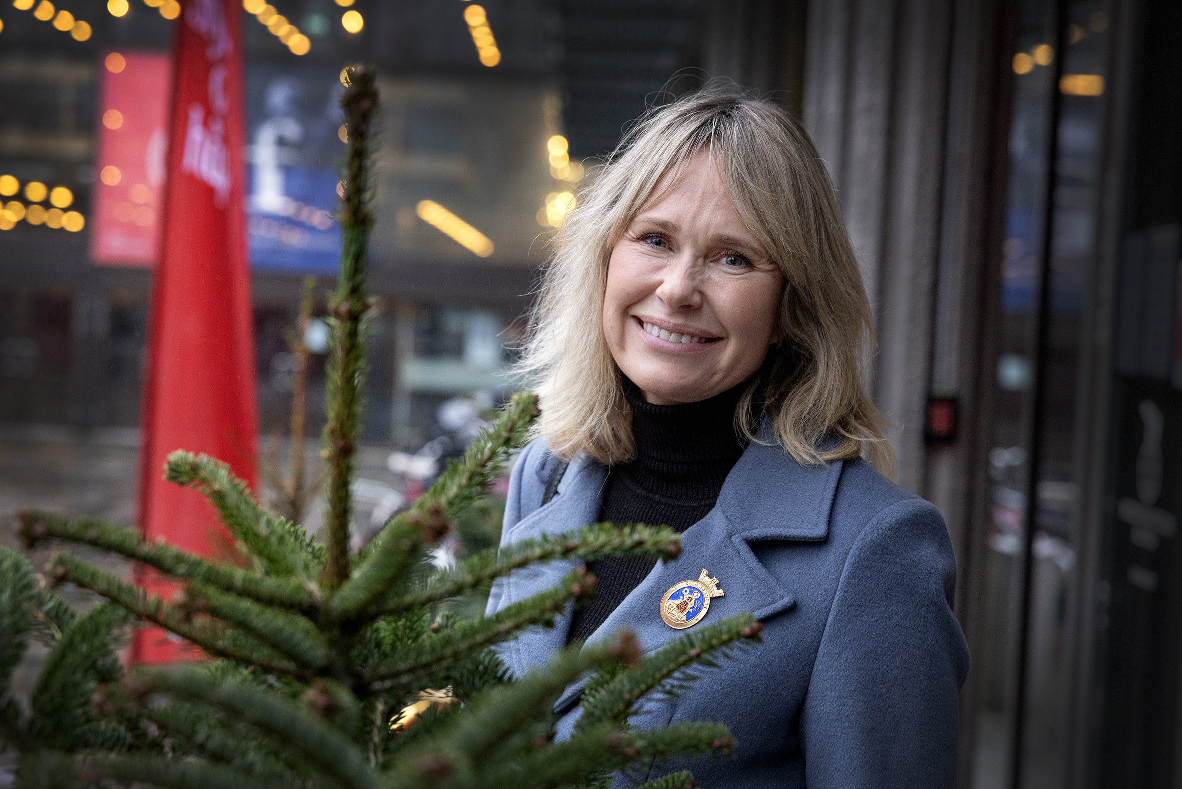Kvinne ser inn i kamera. Står ute, ved siden av et mindre grantre. Treet er ikke pyntet og kun toppen vises. Hun har på seg Oslo kommunes emblem på jakkeslaget.