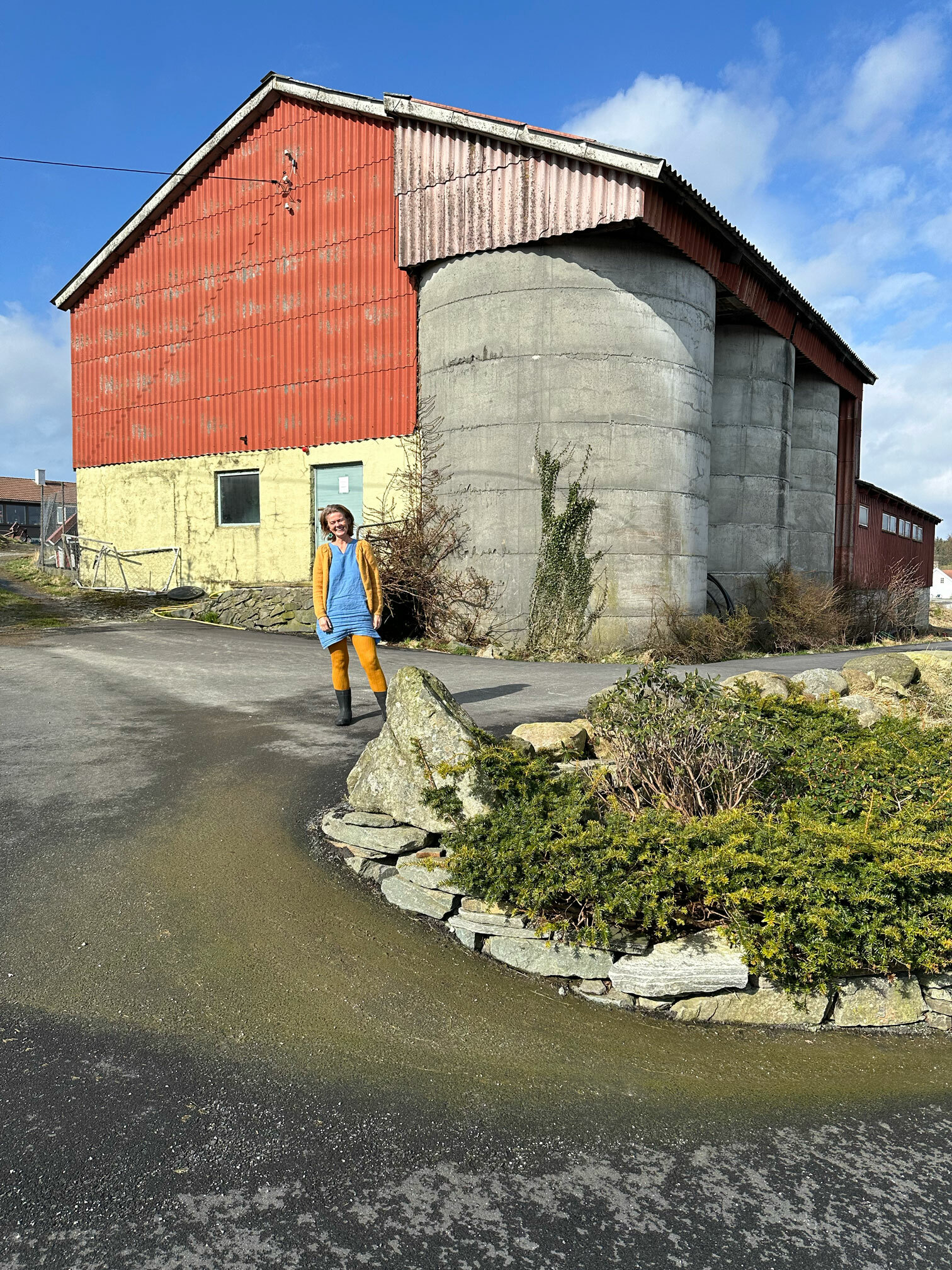 Foto: Kvinne i blå kjole med gul jakke og strømpebukse står i gummistøvler foran veien opp til en driftsbygning på en gård.