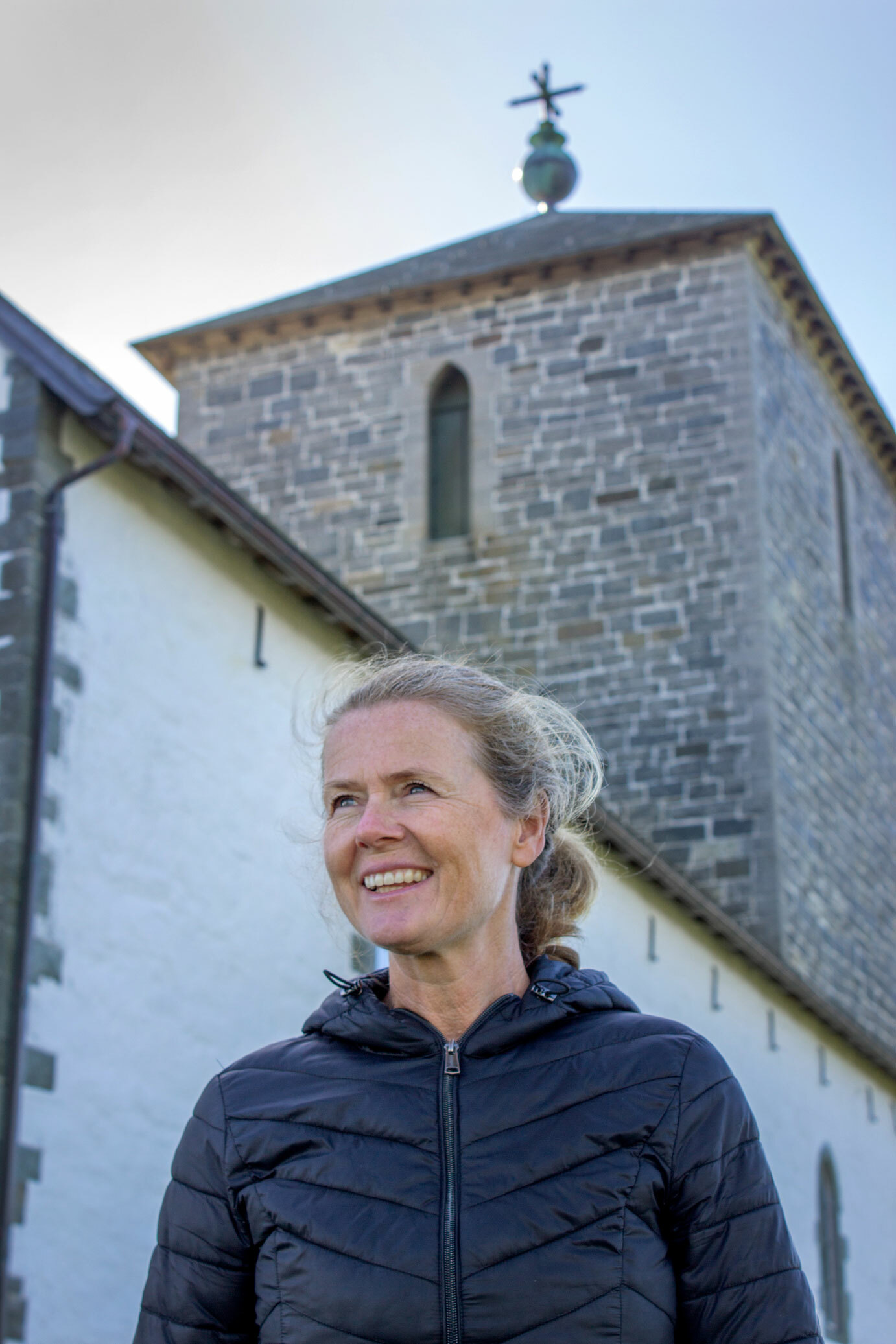 Foto: Kvinne i marineblå boblejakke speider utover. Bak hennes sees spiret på Avaldsnes kirke.