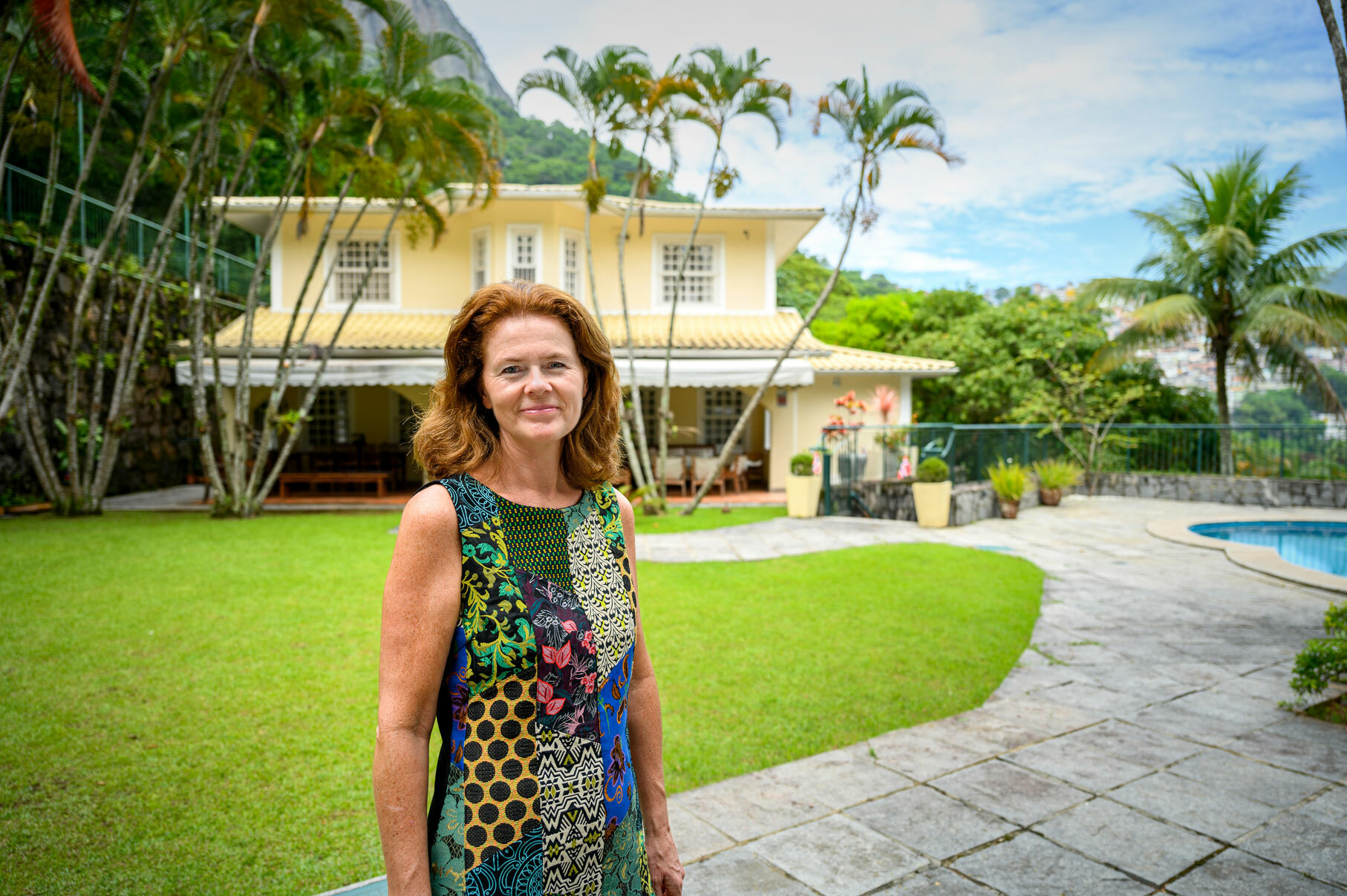 Foto: Kvinne i mønstrete kjole og rødt, halvlangt hår foran et gult murhus med basseng i hagen.