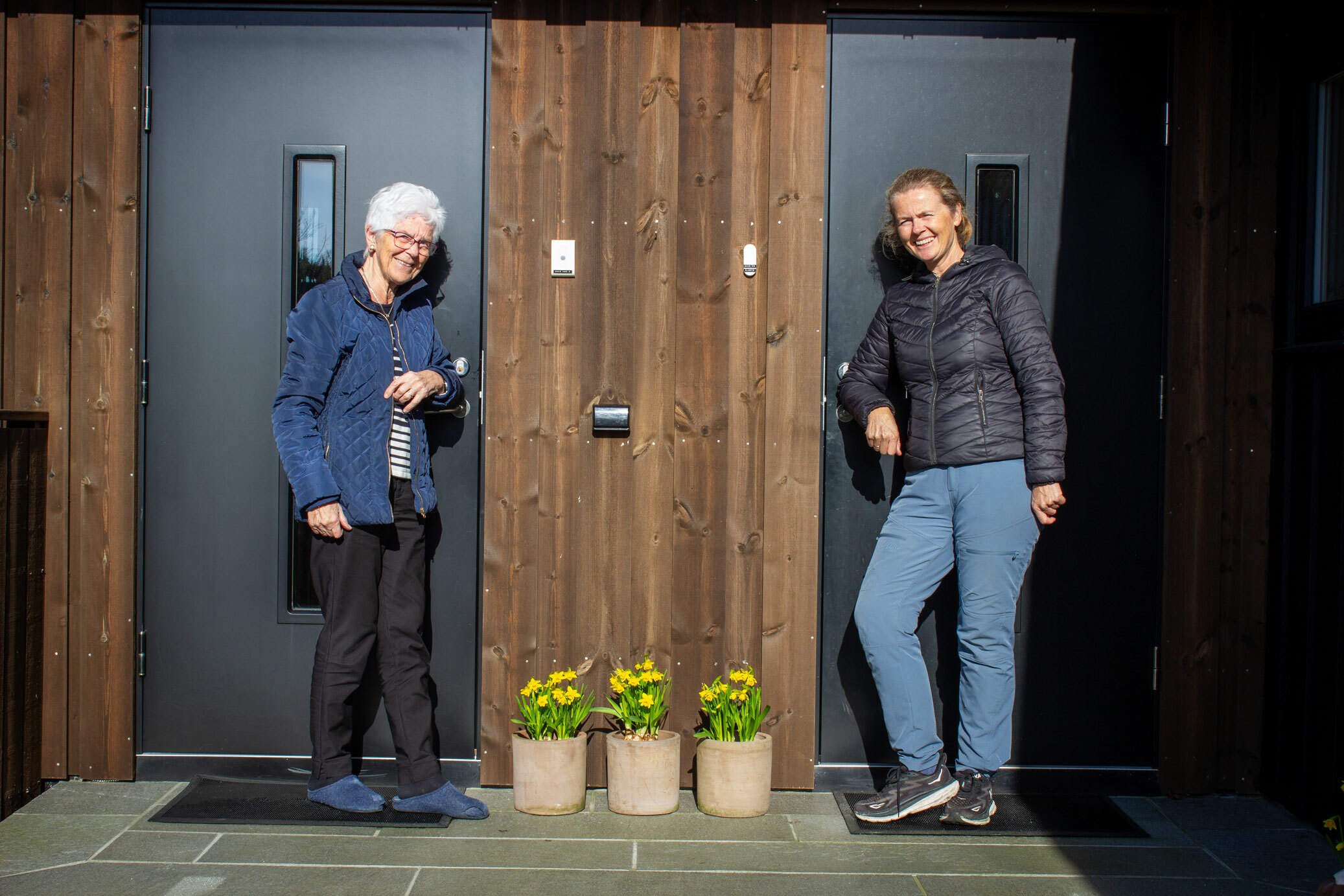Foto: To inngangsdører ved siden av hveramdre. Der står en eldre kvinne utenfor den ene og en middelaldrende i marineblå boblejakke utenfor den andre.