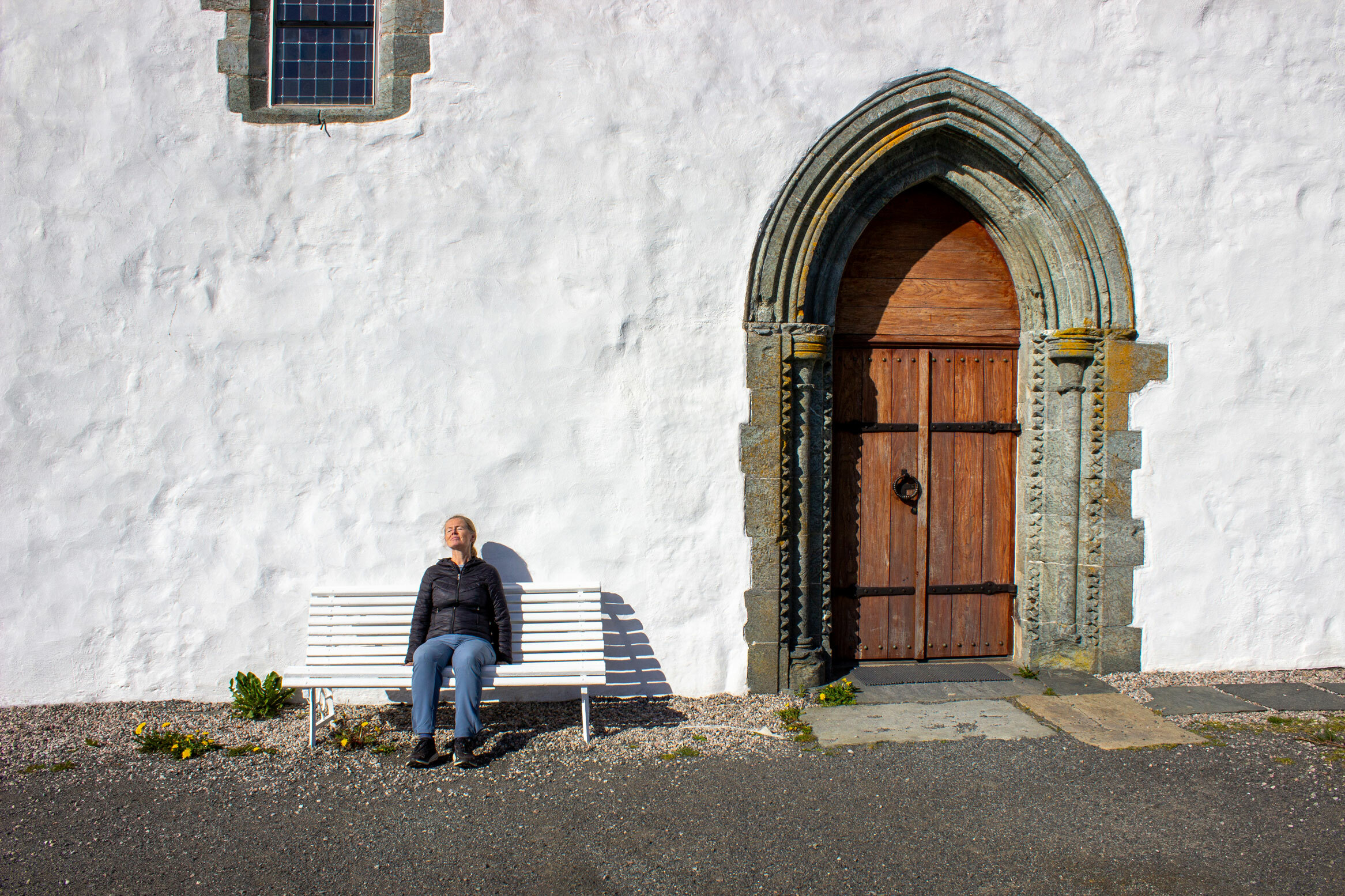 Foto: Kvinne i en marineblå boblejamme sitter på en hvit benk foran en hvitkalket kirkevegg med en stor, buet inngangsdør i tre.