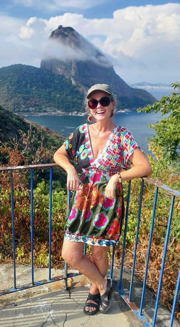 Foto: Kvinne i fargerik kjole og med store solbriller står langs et gjerde med armene oppå. I bakgrunnen sees sjø og en mektig fjelltopp.