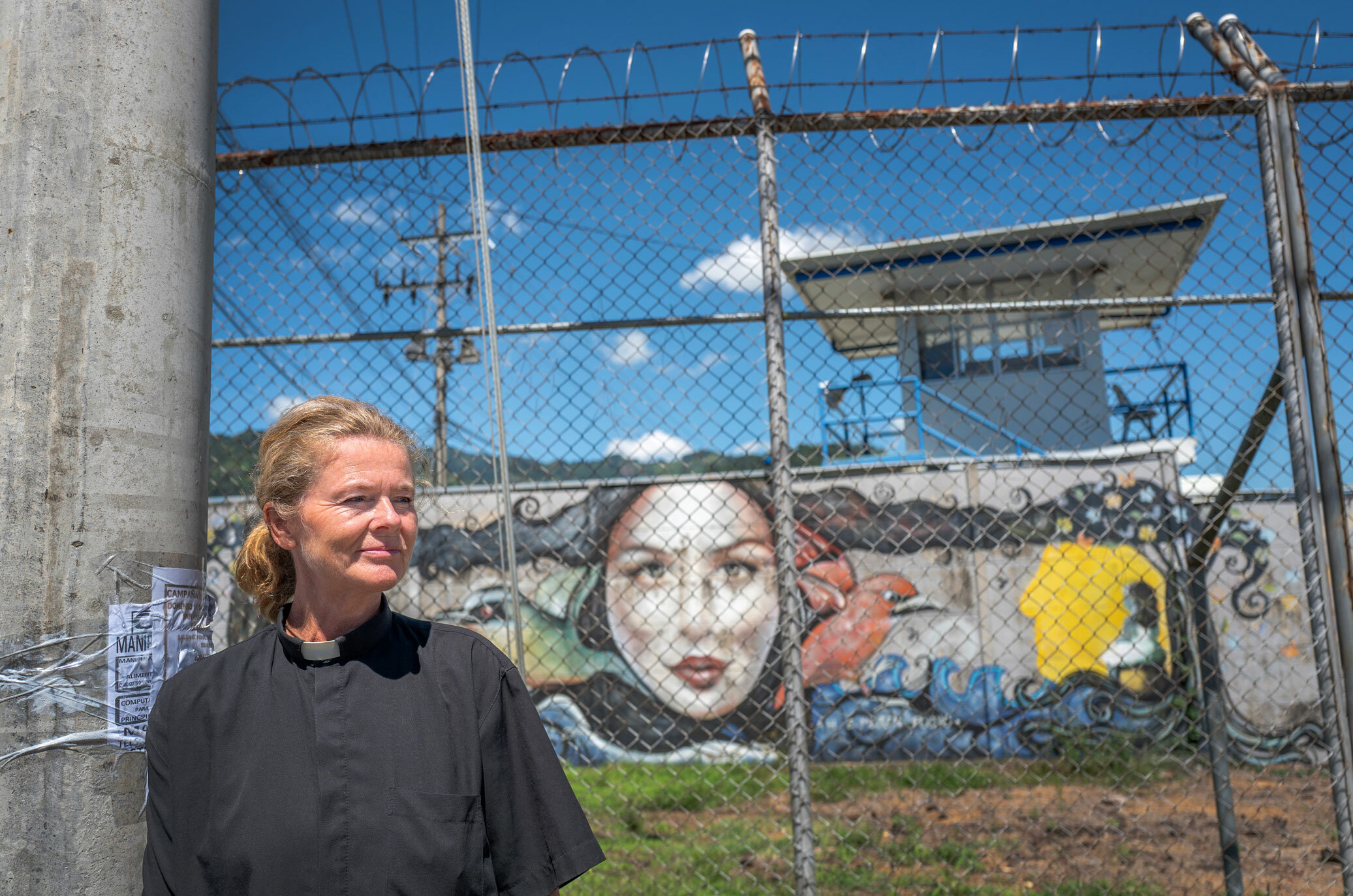 Foto: Kvinne med hvit prestesnipp og svart skjorte står utenfor et høyt nettinggjerde med piggtråd på toppen. Veggen innenfor har grafittitegninger.