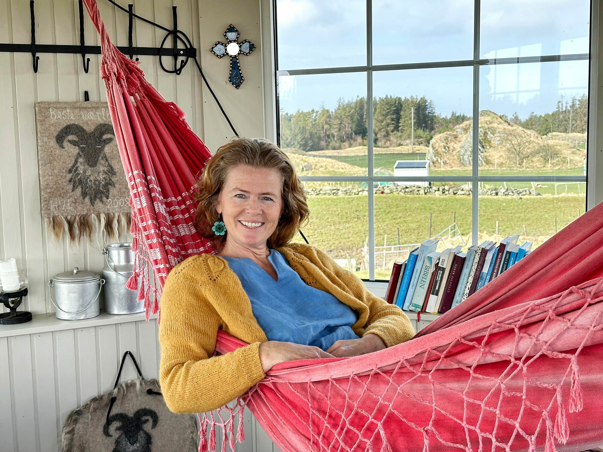 Foto: Kvinne med rødt hår, blå kjole og gul strikkejakke ligger i en rød hengekøye i et lite rom. Utenfor ses beitemarker. I den ene vinduskarmen står flere bøker.