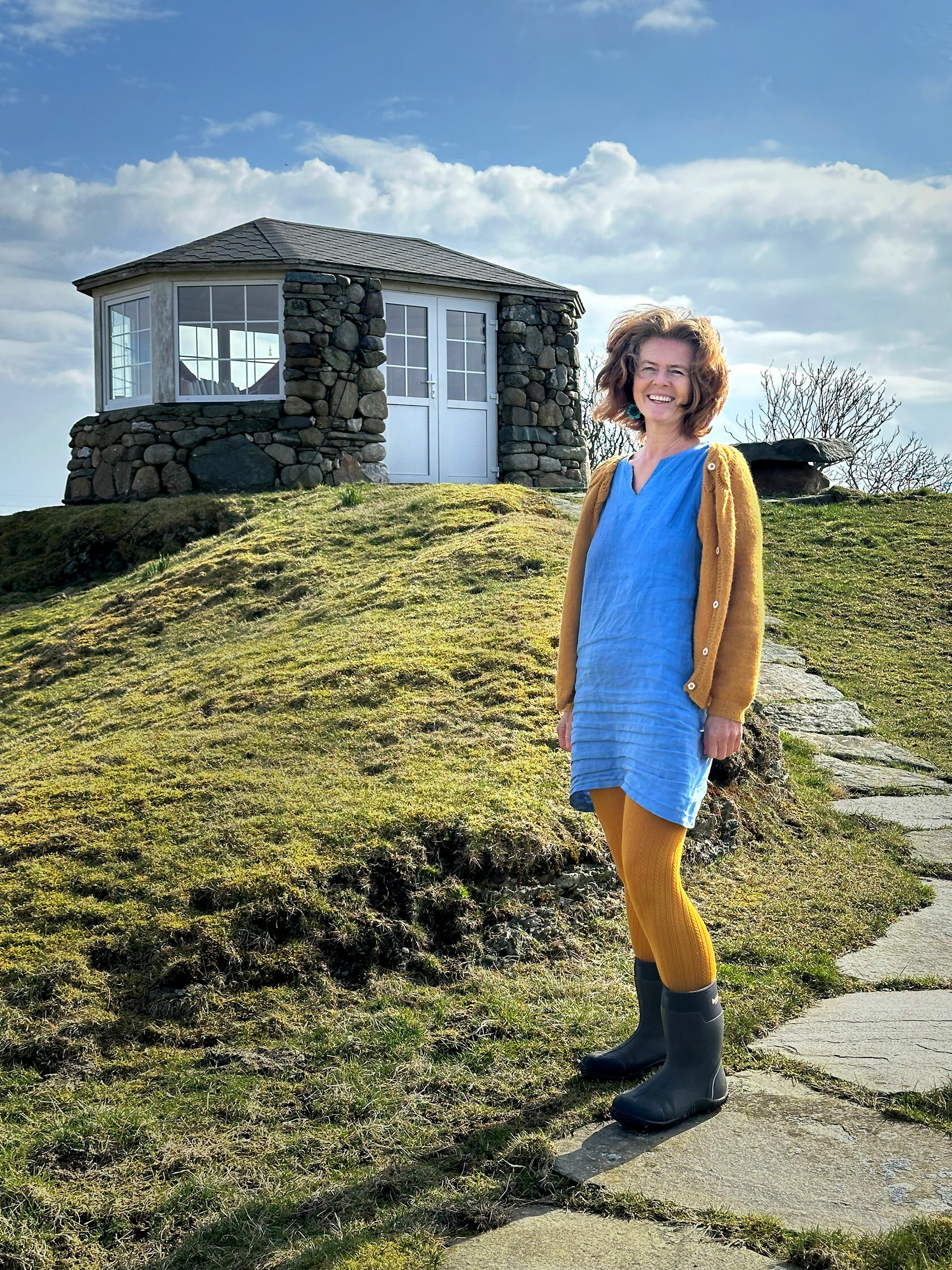 Foto: Kvinne med rødt hår står på en hellebelagt sti opp til en hytte i stein. Hun har på seg blå kjole, gul strikkejakke og strømpebukse og gummistøvler.