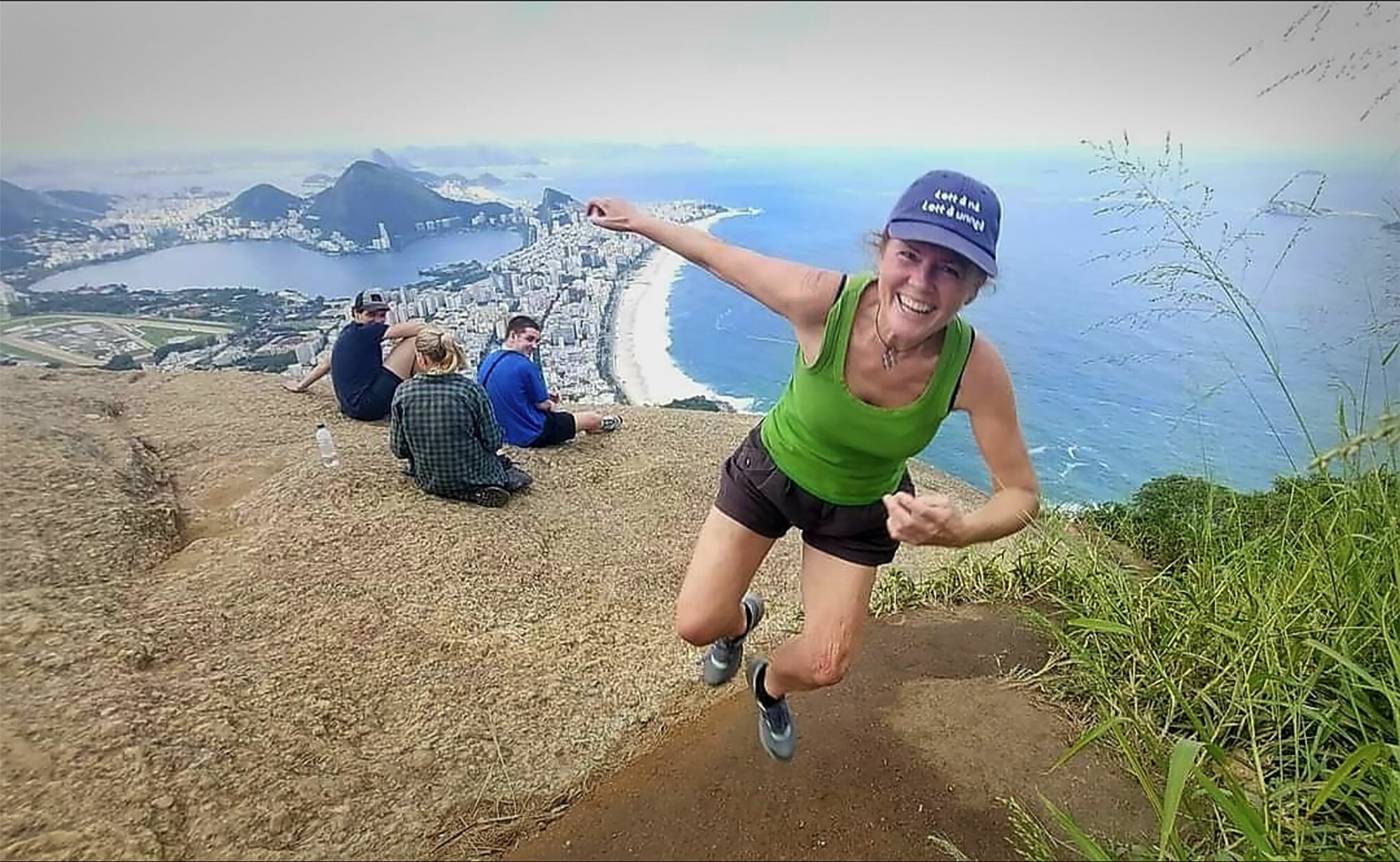 Foto: Kvinne i grønn topp, shorts og caps hopper og ler. I bakgrunnen sees en hvit strand og fjell.