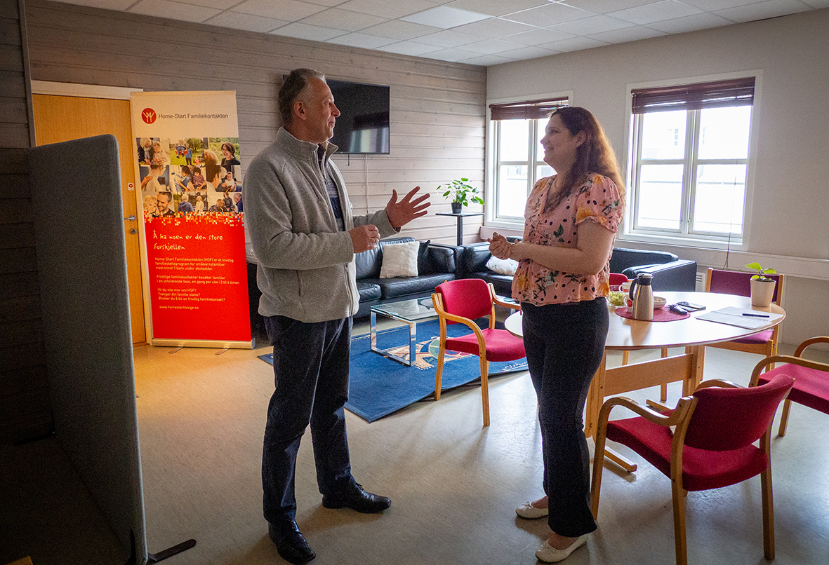 Mann i 60-årene står og snakker med en kvinne med mørkt, krøllete hår. De står i et rom med to sofaer og mange stoler rundt et bord.