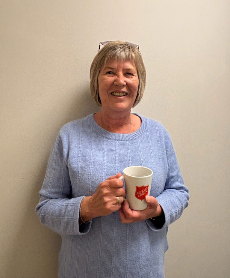 Kvinne i 60-årene med grått hår og blå genser står med en kaffekopp i hånden. Den har rød logo fra Frelsesarmeen.