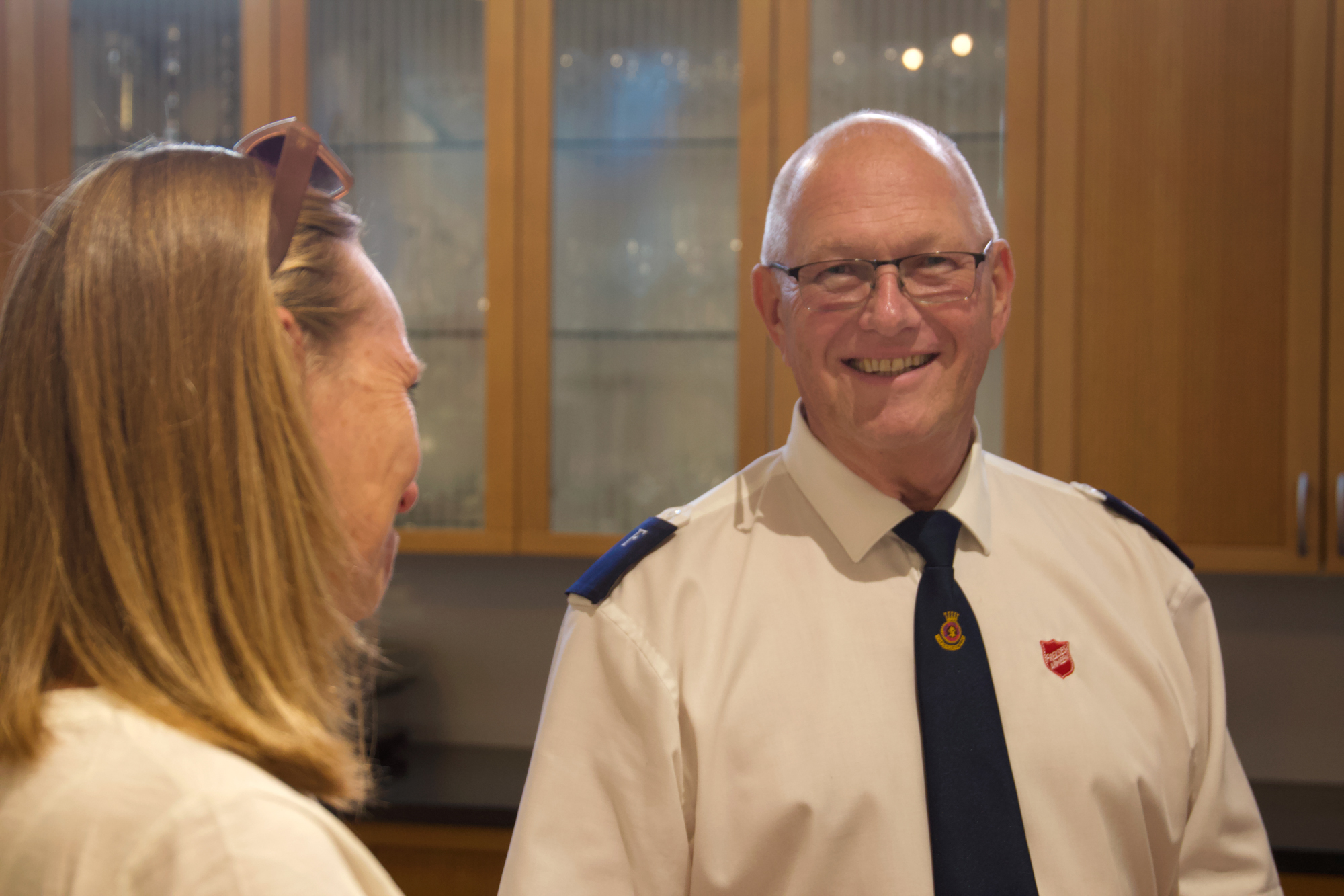Foto: Dame i 50-årene med blondt hår snakker med en mann i hvit uniformskjorte fra Frelsesarmeen og blått slips. Han er blank på hodet og bruker briller. Begge smiler.