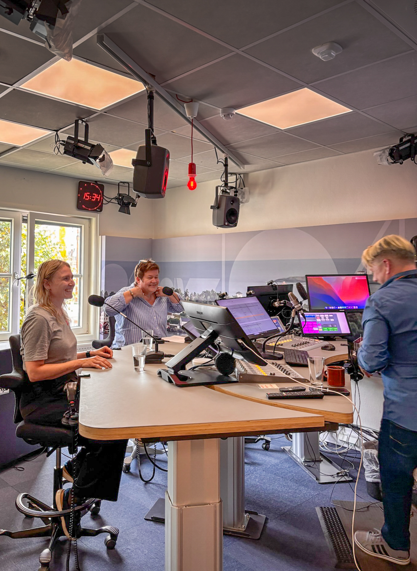 Foto: To kvinner sitter i et lydstudio sammen med en stående, mannlig programleder.