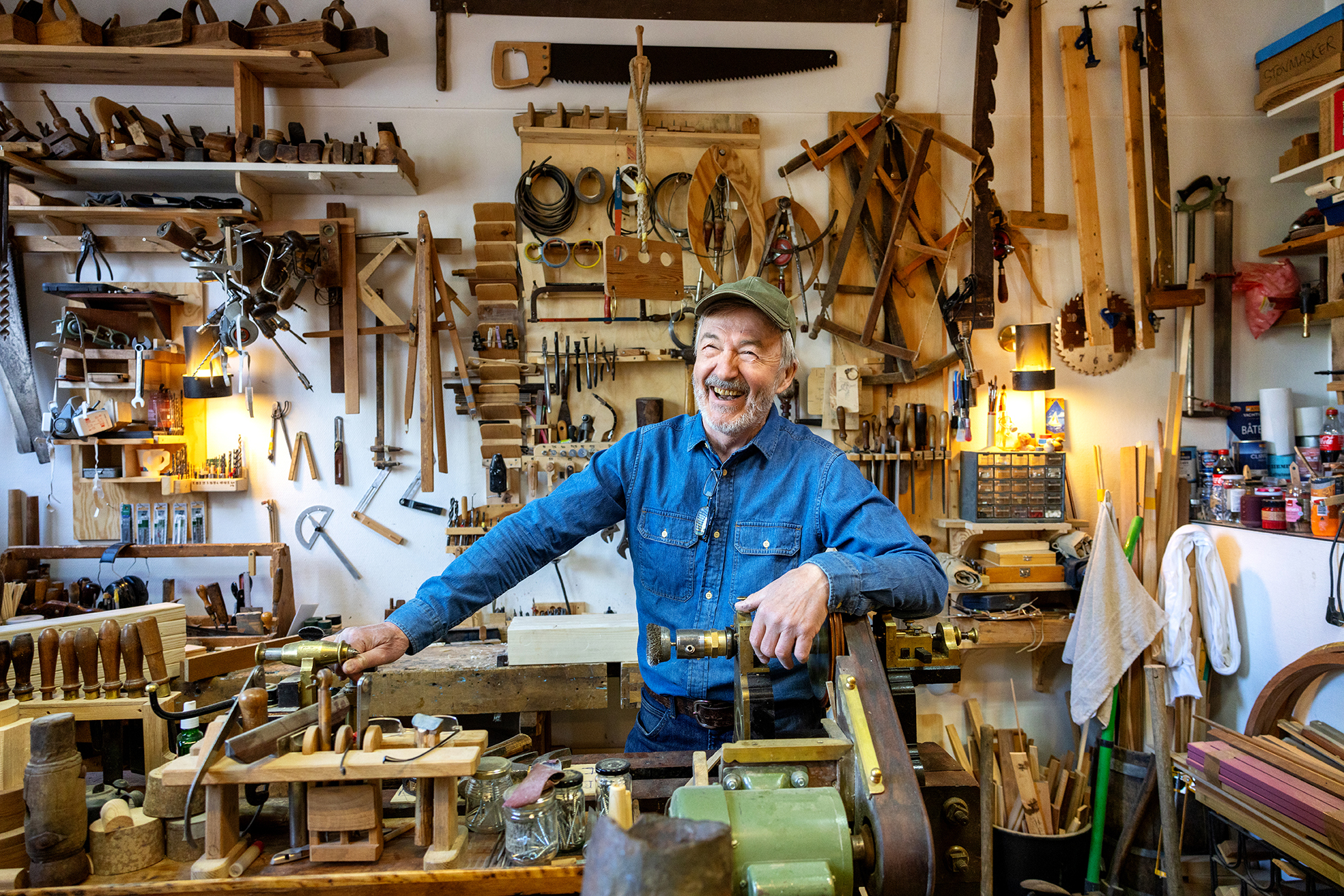 Foto: Eldre mann i blå kjeledress og grønn caps står i et snekkerverksted. Veggen bak er full av redskaper.