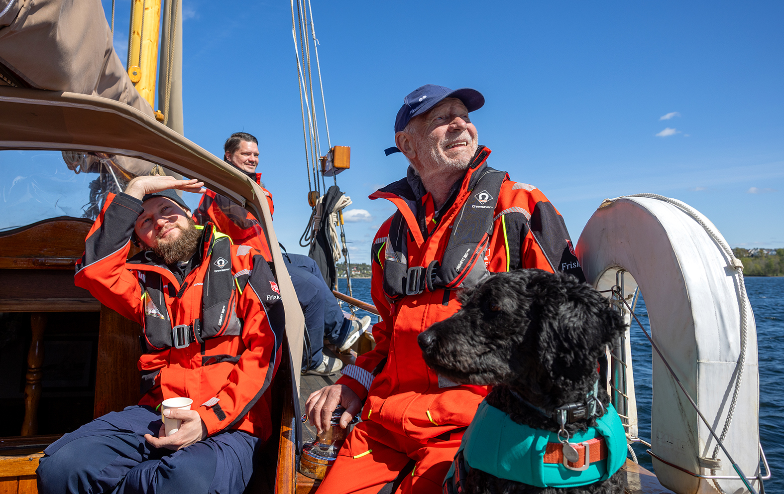 Foto: Tre menn i seilertøy står og sitter på dekk sammen med en svart hund med grønn redningsvest.