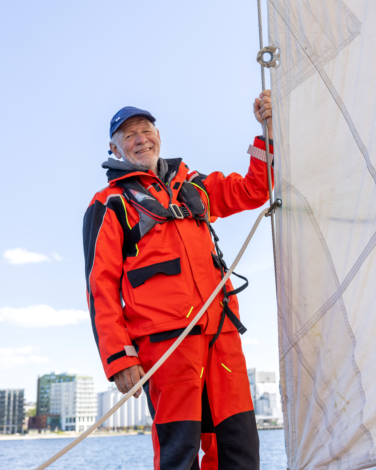 Foto: Godt voksen mann med grått skjegg og røde klær står ved seilet. I bakgrunnen sees kontorbygg ved vannkanten i Moss.
