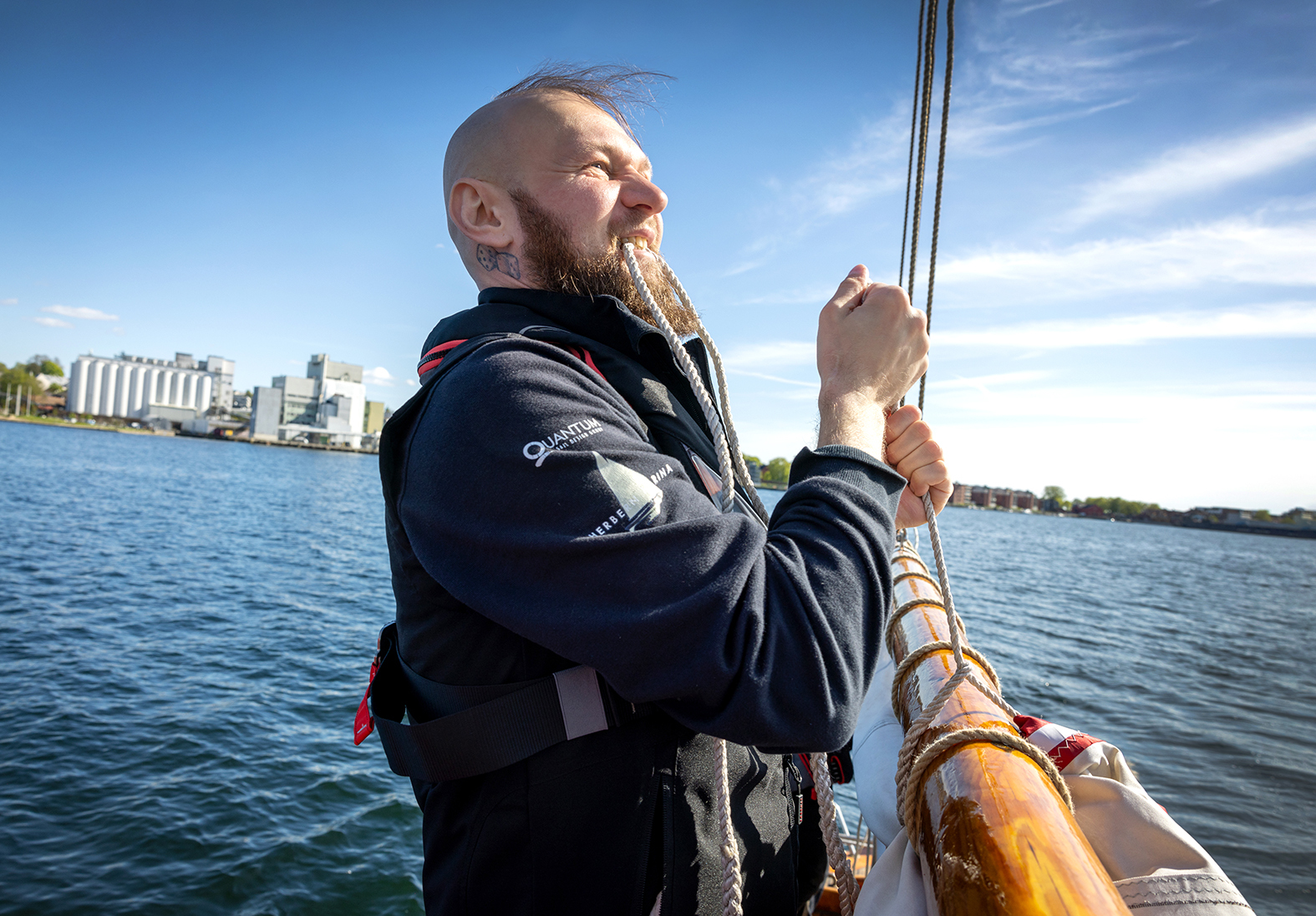 Foto: Mann i mørkt seilerutstyr drar i et tau på en seilbåt. Han holder i ene enden av det med tennene. Han har skjegg og er nesten glattbarbert på hodet, det stikke bare litt hår opp fra toppen.