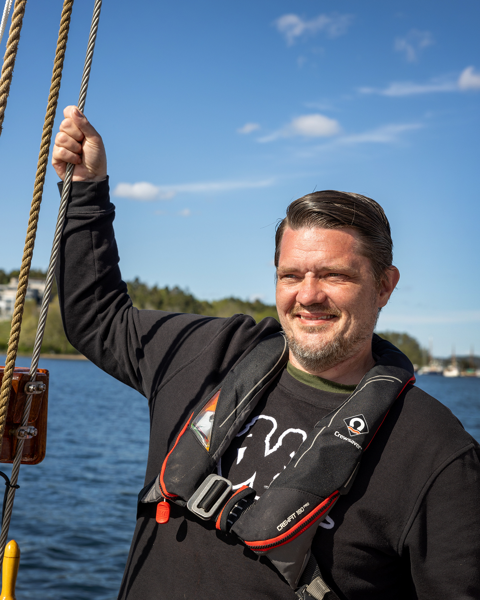 Foto: Mann i blå genser og redningsvest står og holder seg fast i et tau. Han kikker utover.