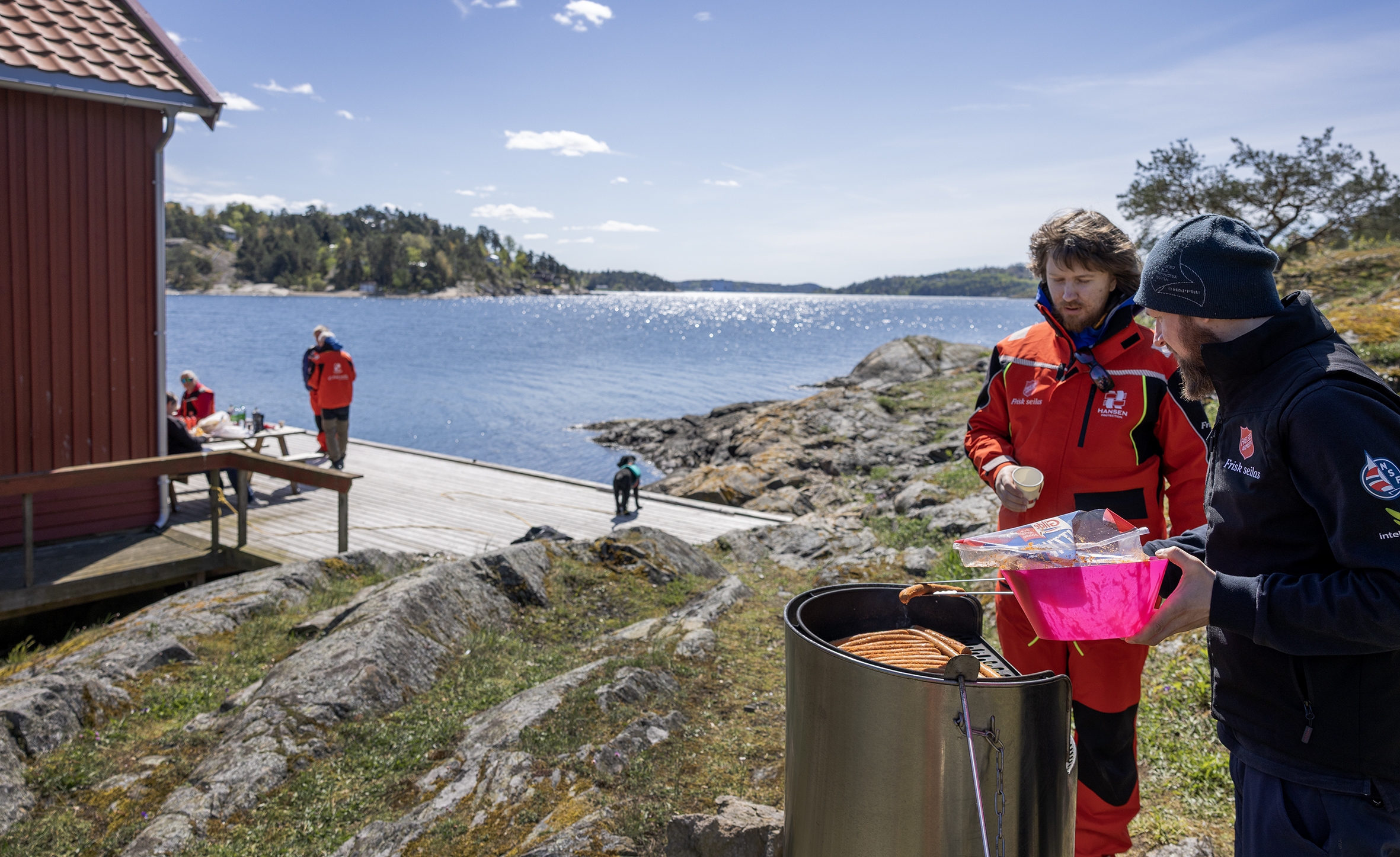 Foto: To menn står ved en grill på en holme. Havet glitrer i bakgrunnen og det er nesten blå himmel, kun et par hvite skydotter.