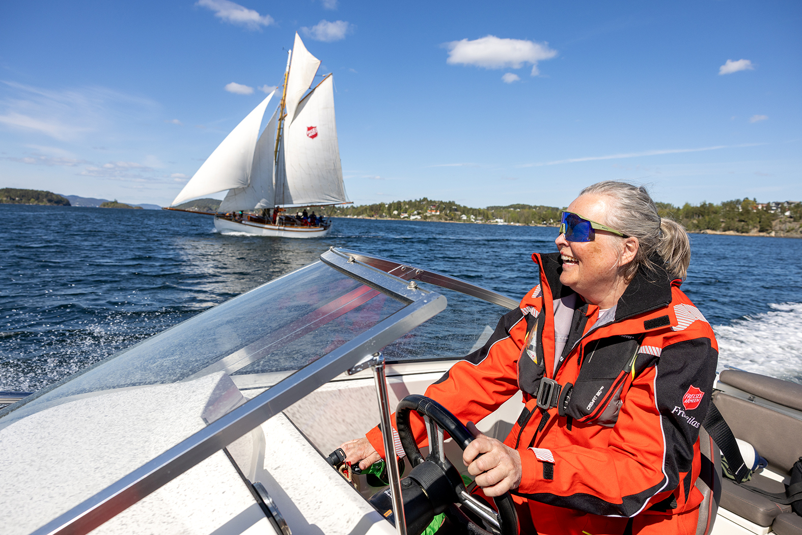 Foto: Gråblond kvinne med håret dratt bak i en hestehale smiler mens hun styrer en speedbåt. Hun har på seg "raske briller"og rød seilerjakke. I bakgrunnen sees seilbåten med alt av seil oppe.