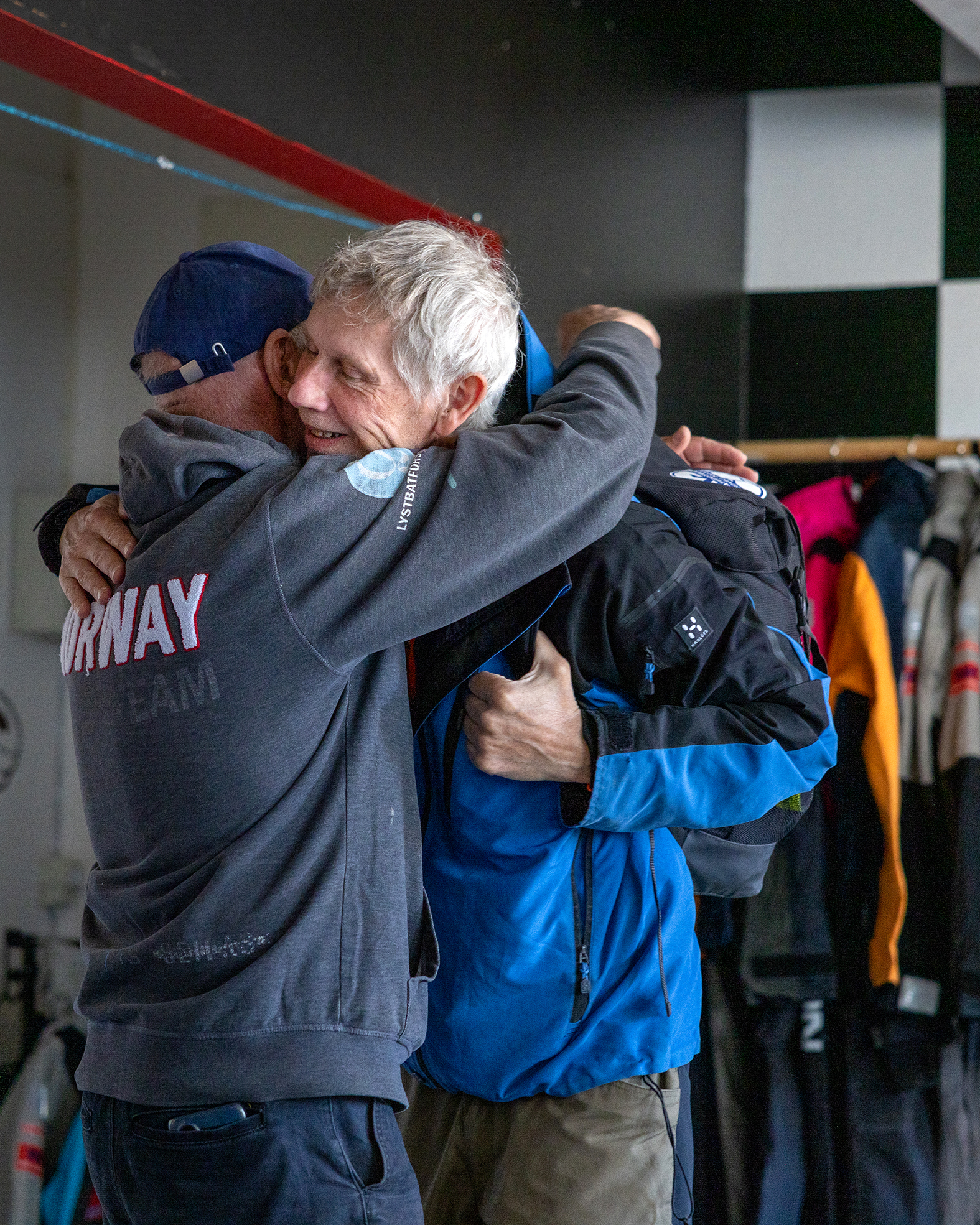 Foto: To menn som gir hverandre en klem inne i et rom der det henger alt av seilerutstyr på rekke og rad.