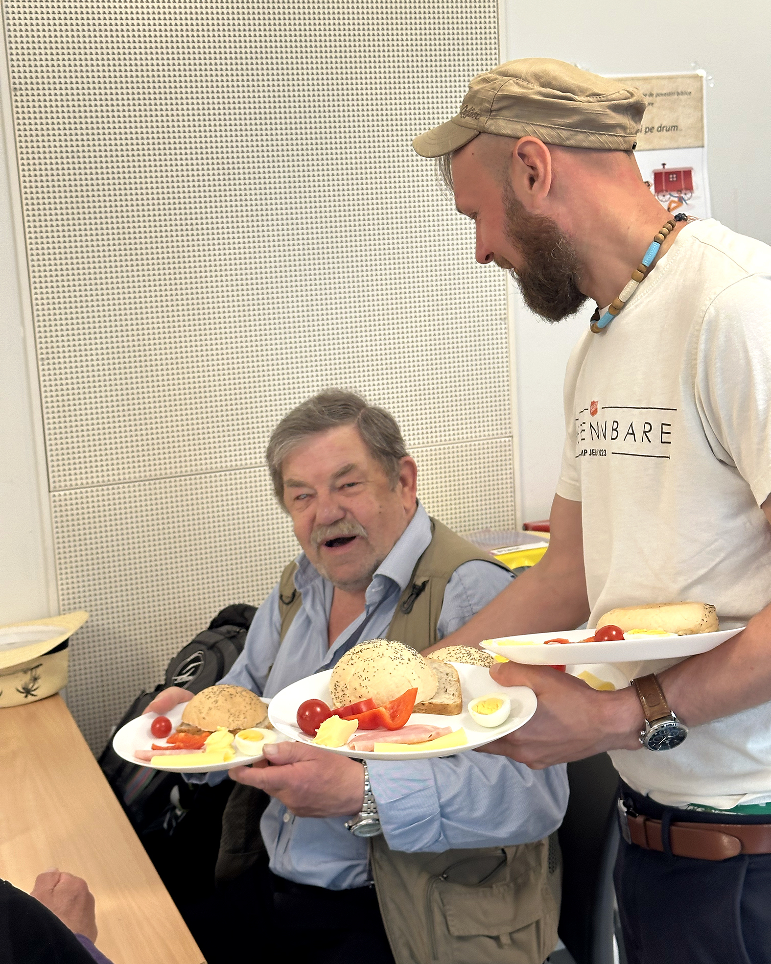 Foto: Mann i hvit T-skjorte og caps serverer mat til en mann med blå skjorte og kaki vest. Han er blind på et øye.