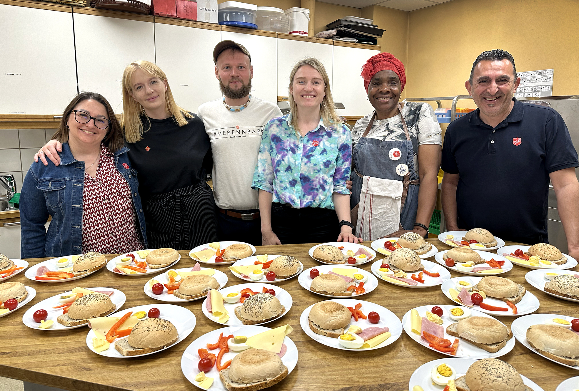 Foto: Fire kvinner og to menn står på rekke inne på et hvitt kjøkken med gule vegger. Foran på bordet ligger det mat på mange fat.