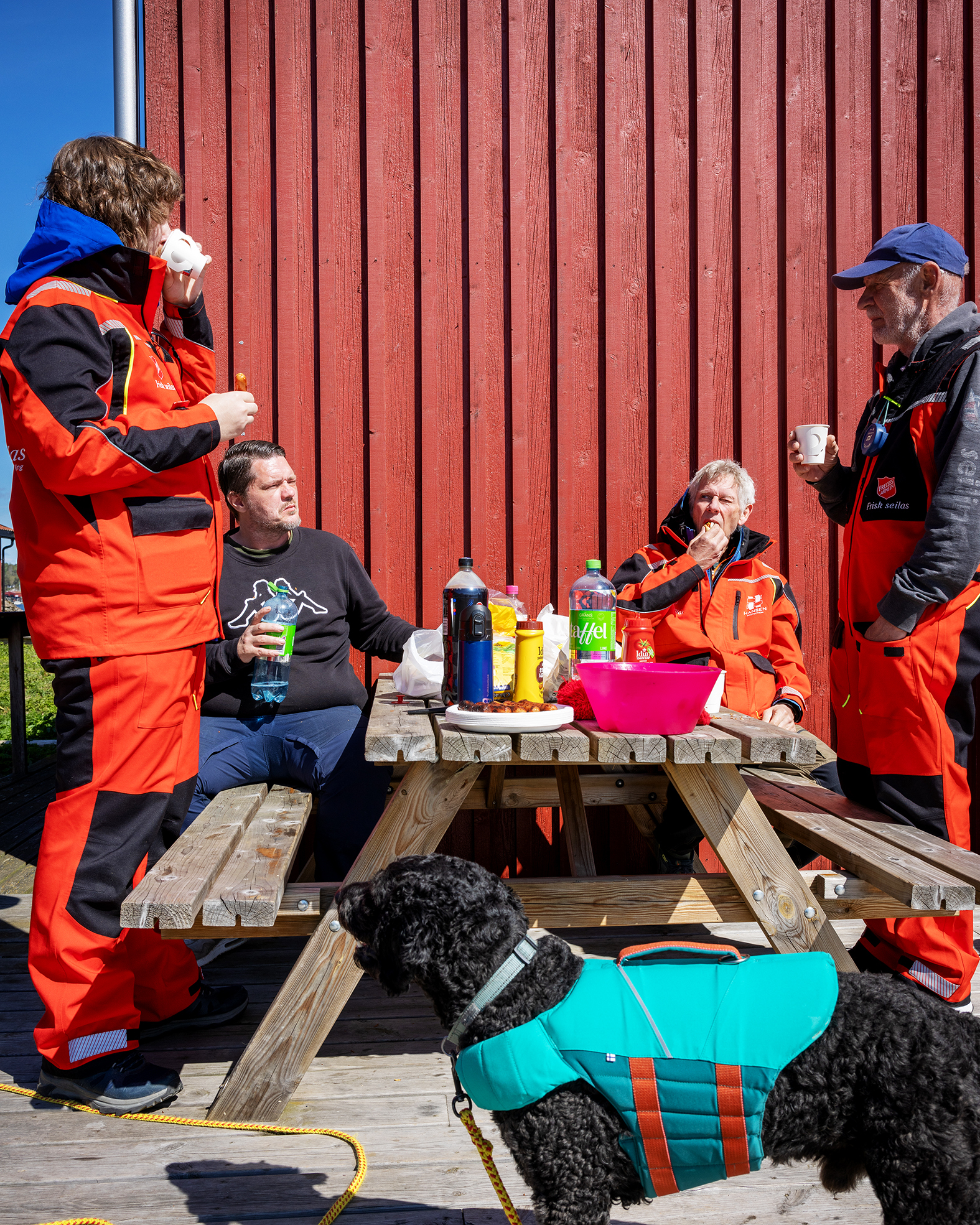 Foto: Fire menn står og sitter ved et utendørs trebord med fastmonterte benker på to av sidene. De sitter i sola, i ly for vinden foran en rødmalt vegg. Foran dem står en svart hund med grønn regningsvest. Det er mat og drikke på bordet.