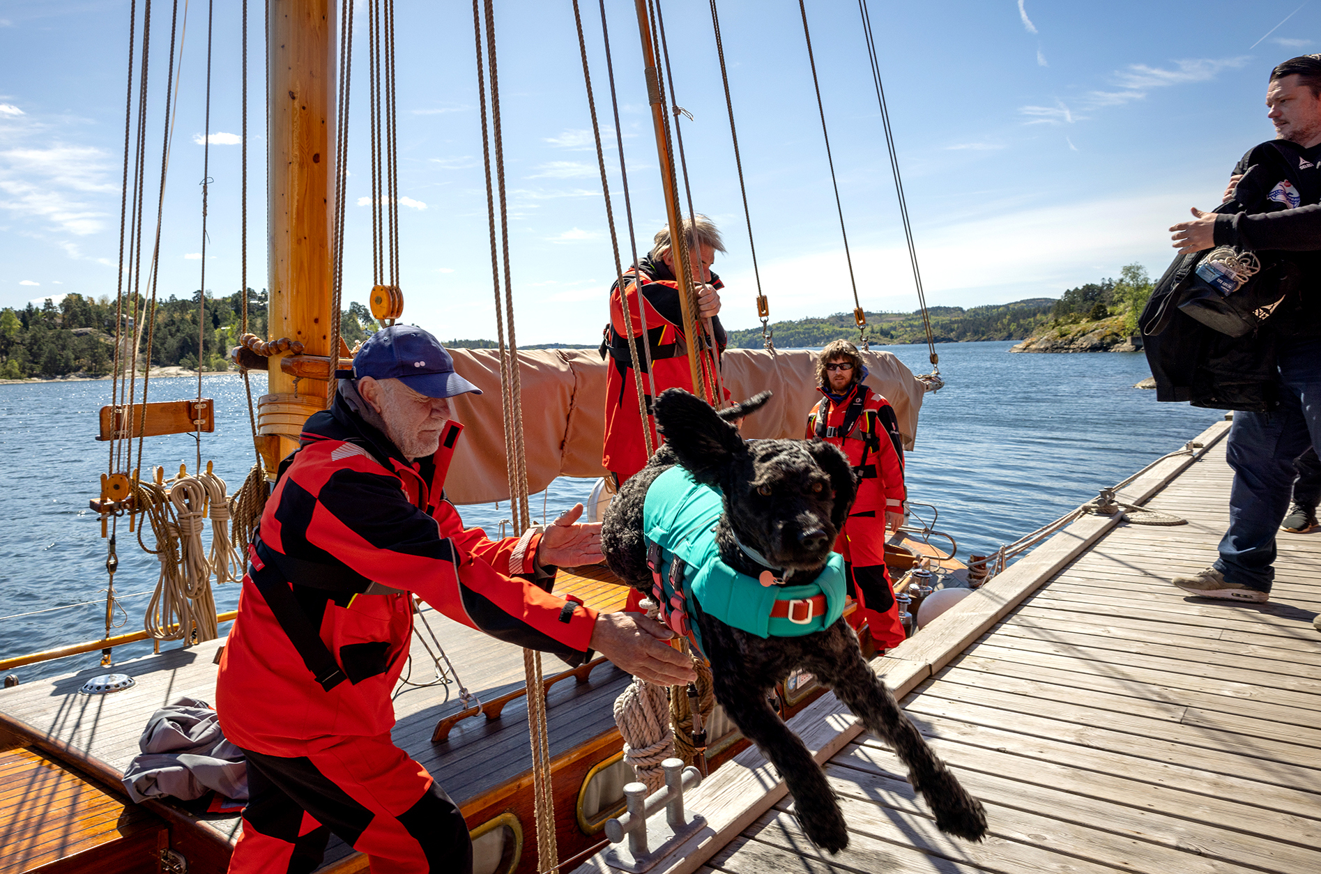Foto: Båt som har lagt til kai. Mannskapet står delvis på kaien og på båten. En svart hund er i svevet mellom båten og kaien.